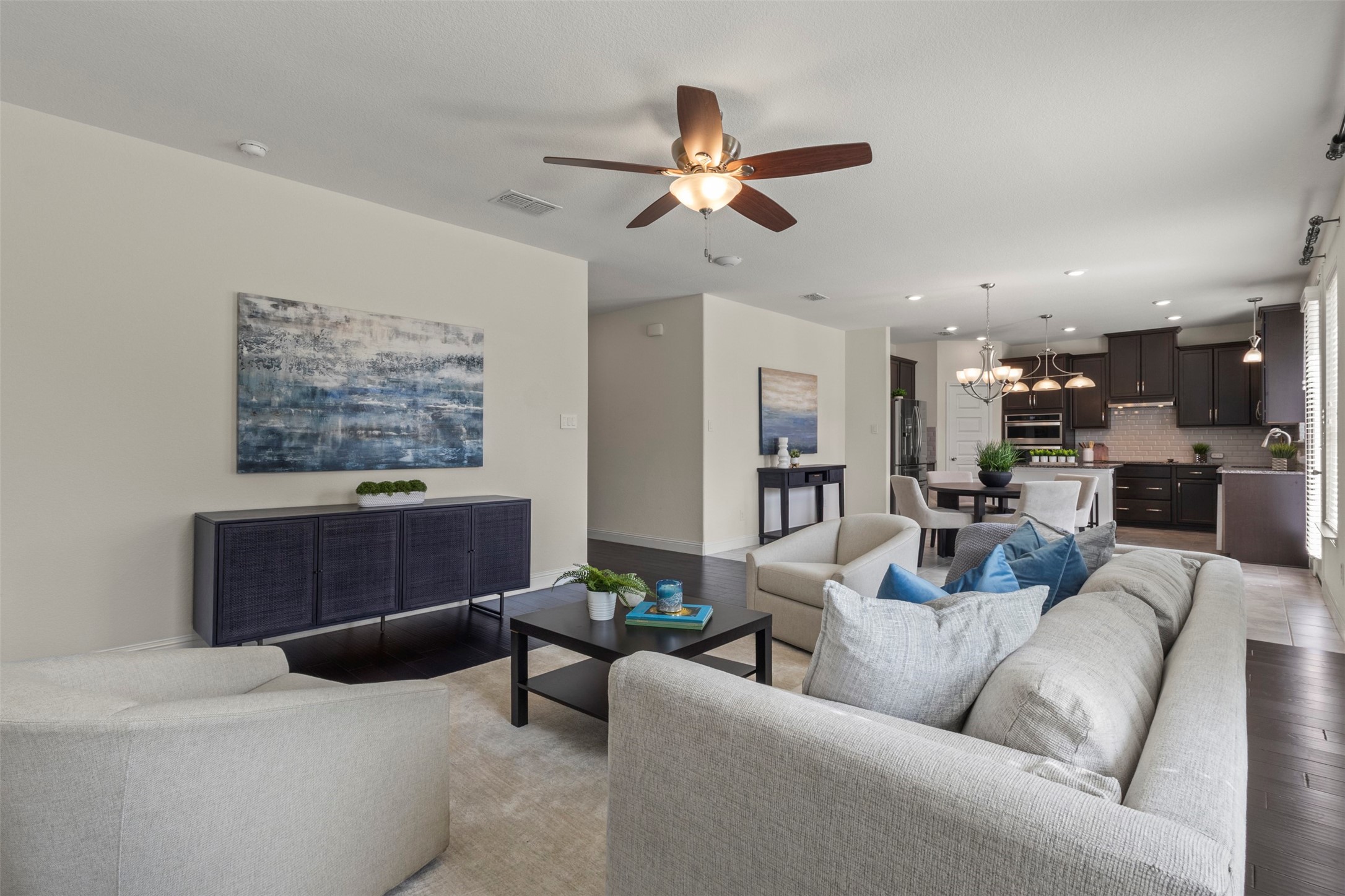Open-plan living room with pale sofas, blue artwork, and a dining area adjoining a dark-cabinet kitchen.