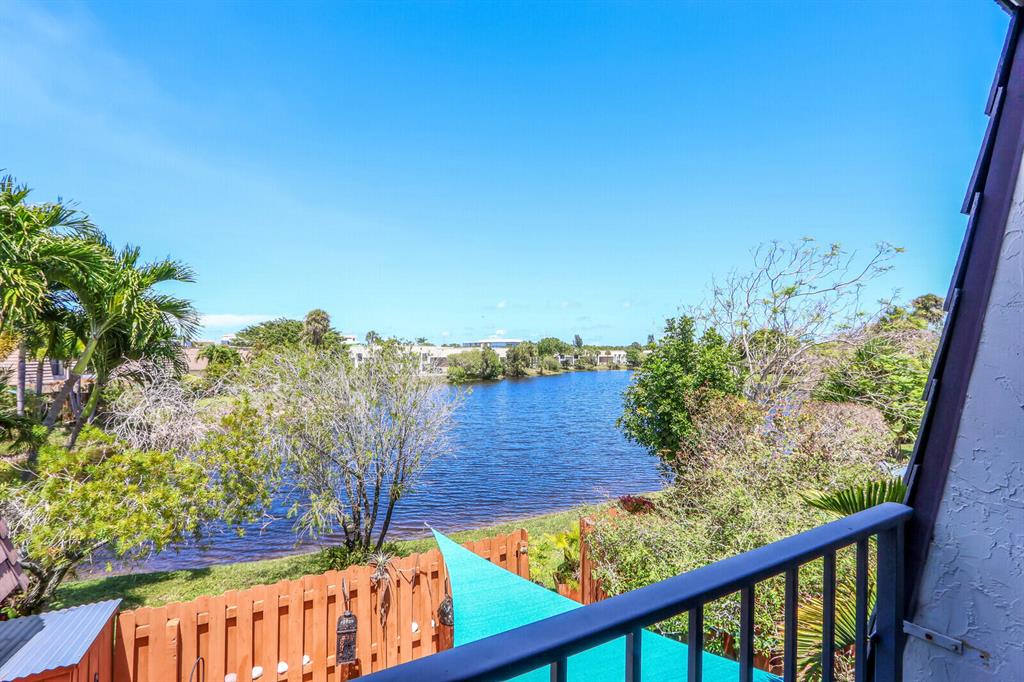 Balcony view across a fenced backyard to a lake, with tropical landscaping and a turquoise shade canopy.