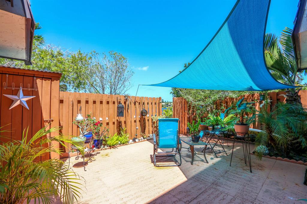 Fenced patio with a blue shade sail, potted plants, and outdoor seating.