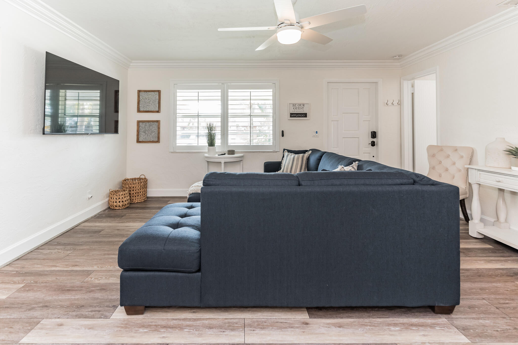 Living room with a dark blue sectional, plantation shutters, and wood-look tile flooring.