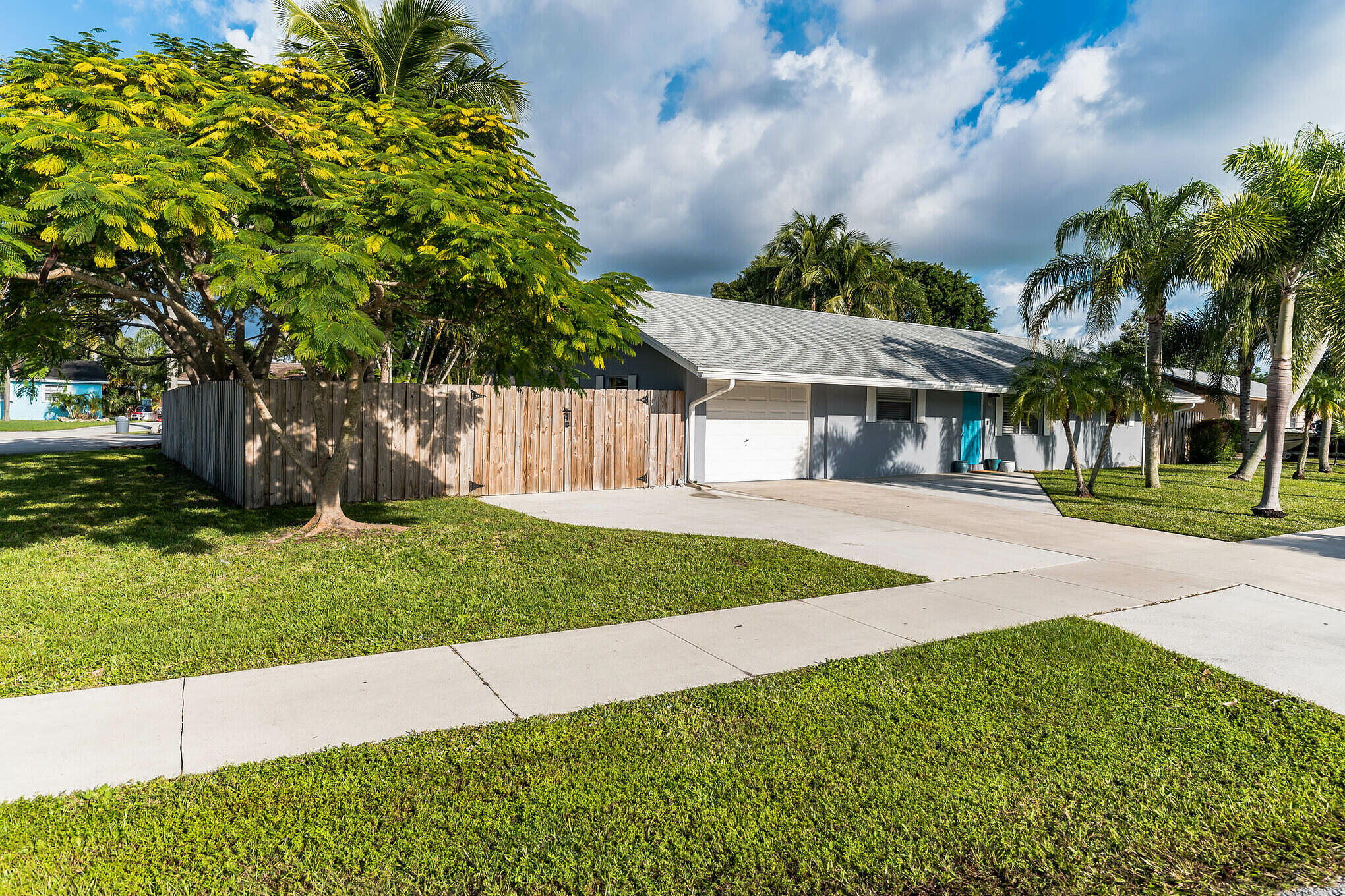 Single-story gray home with a white garage, broad driveway, wooden fence, and tropical landscaping.