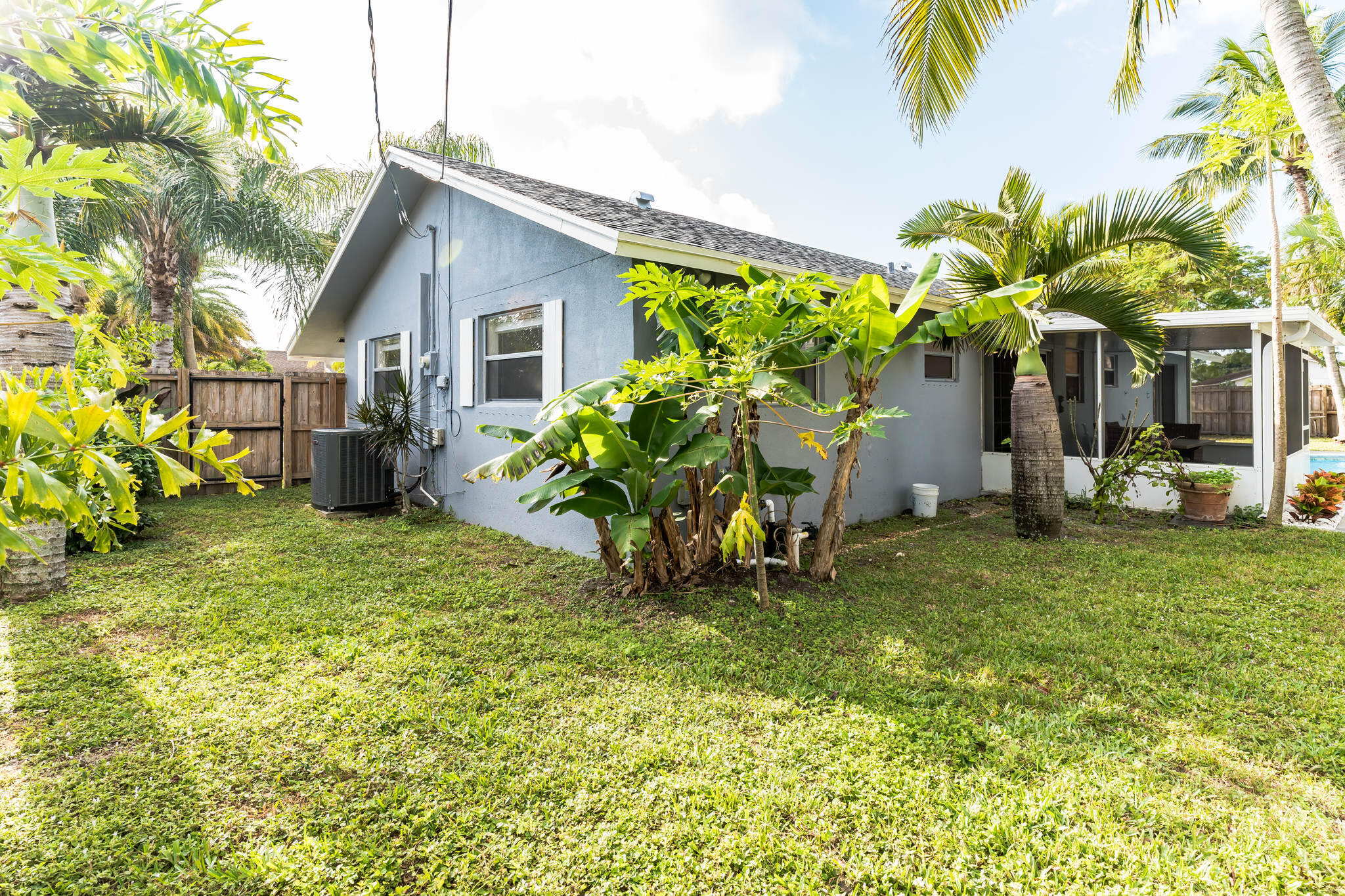 Blue-gray home’s rear exterior with screened patio, grassy yard, and lush tropical plants.