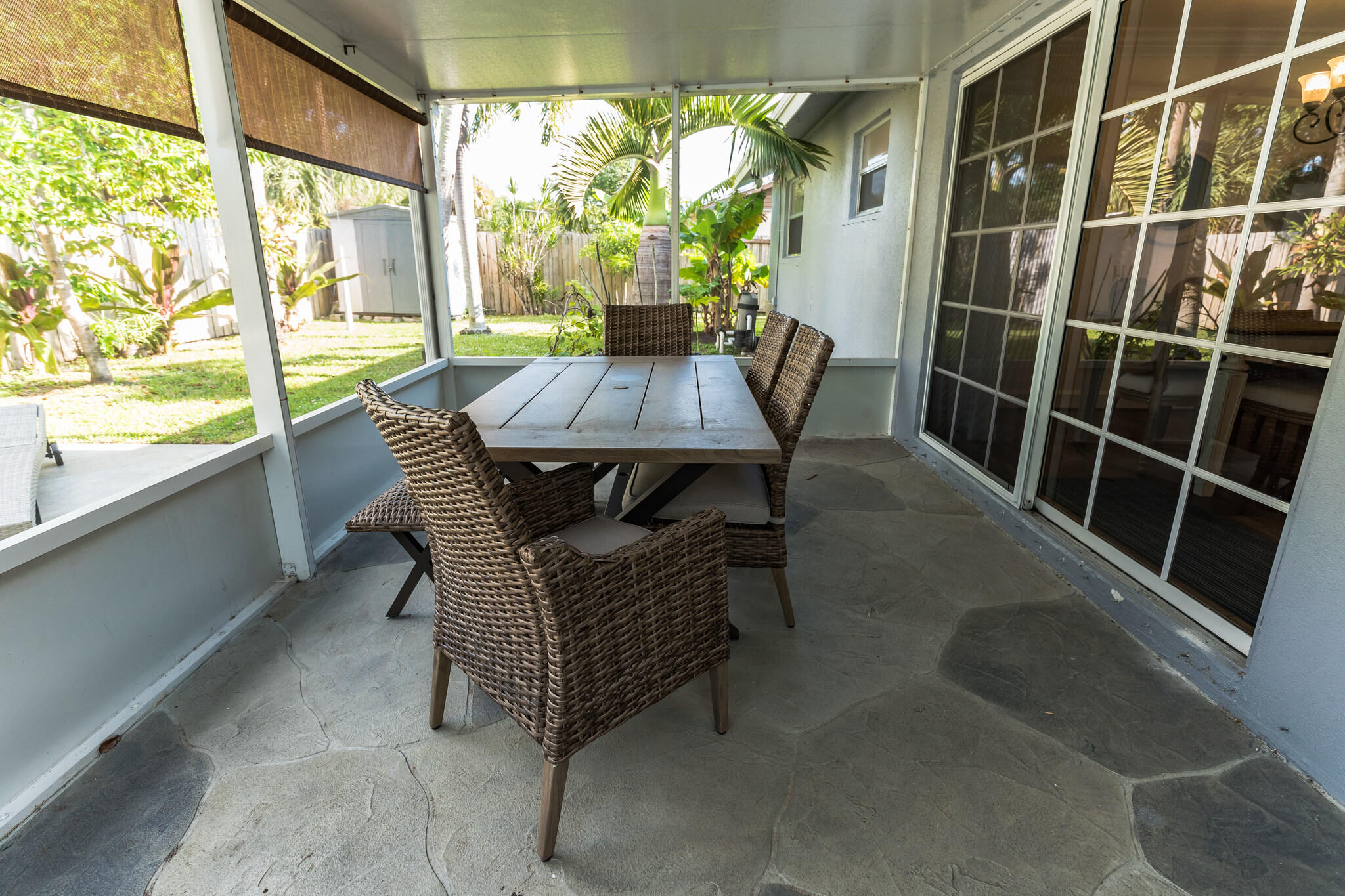 Screened patio with wicker dining set, stone flooring, and a view of the tropical backyard.