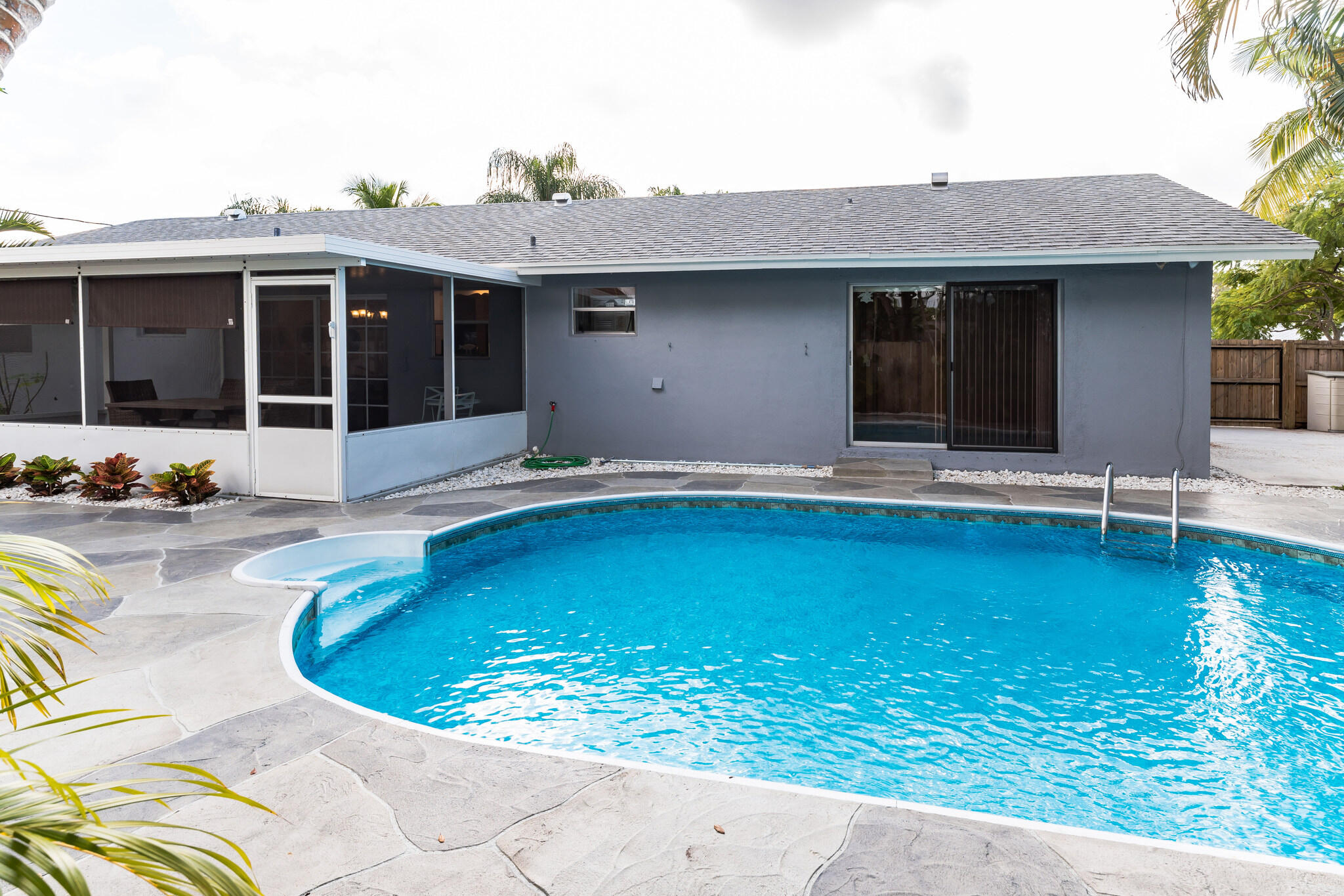 Back of a gray home with a screened patio behind a curved swimming pool and stone deck.
