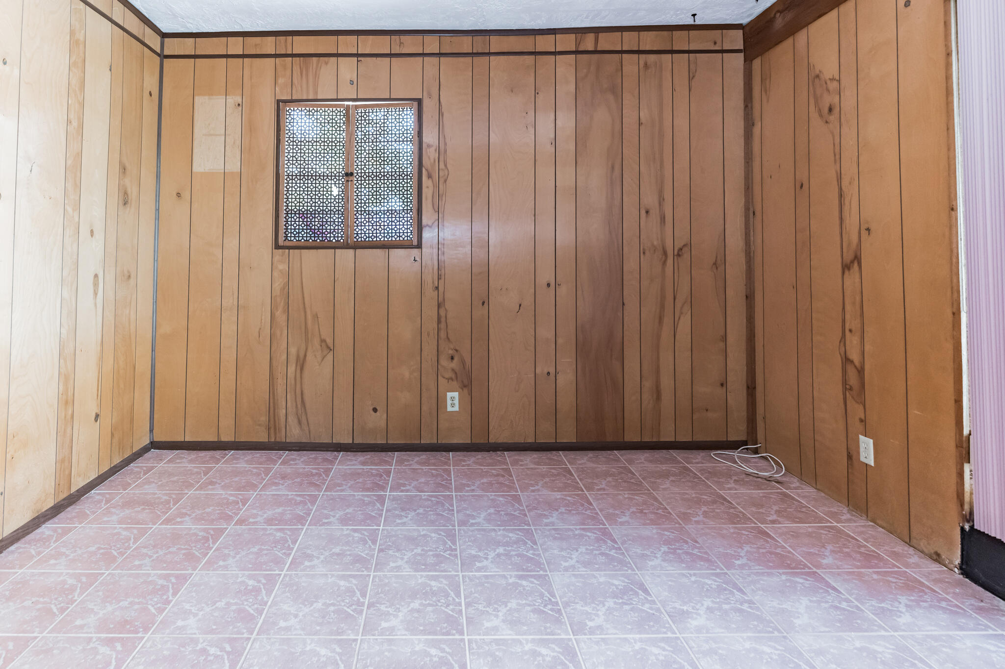 Empty room with wood-paneled walls, pink tile flooring, and a small patterned window.
