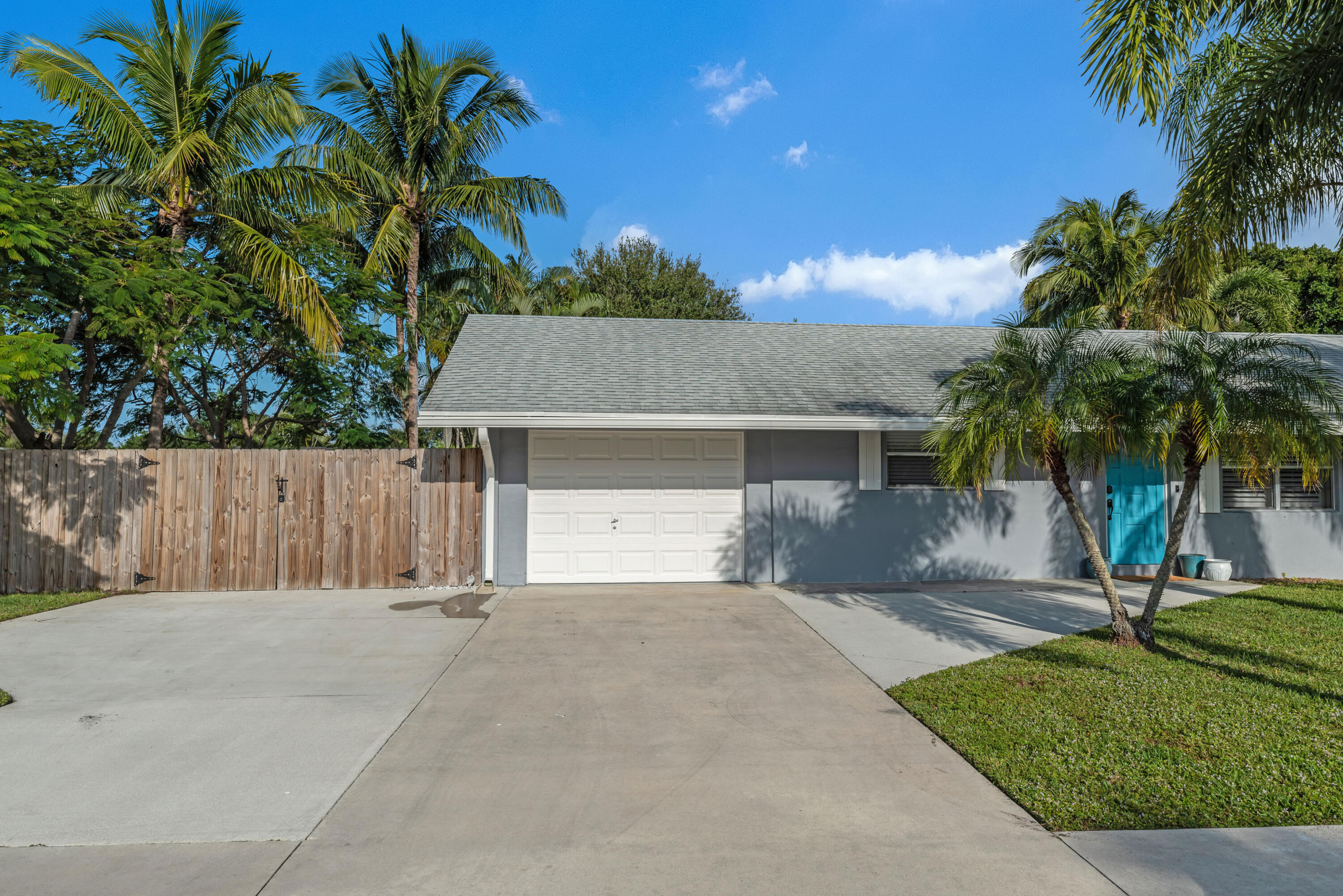Gray single-story home with a turquoise front door, white garage, broad driveway, and palm trees.