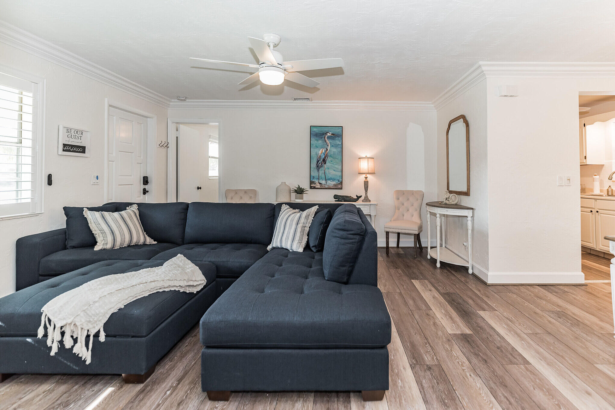 Staged living room with a dark blue sectional, wood-look floors, and an adjoining kitchen.