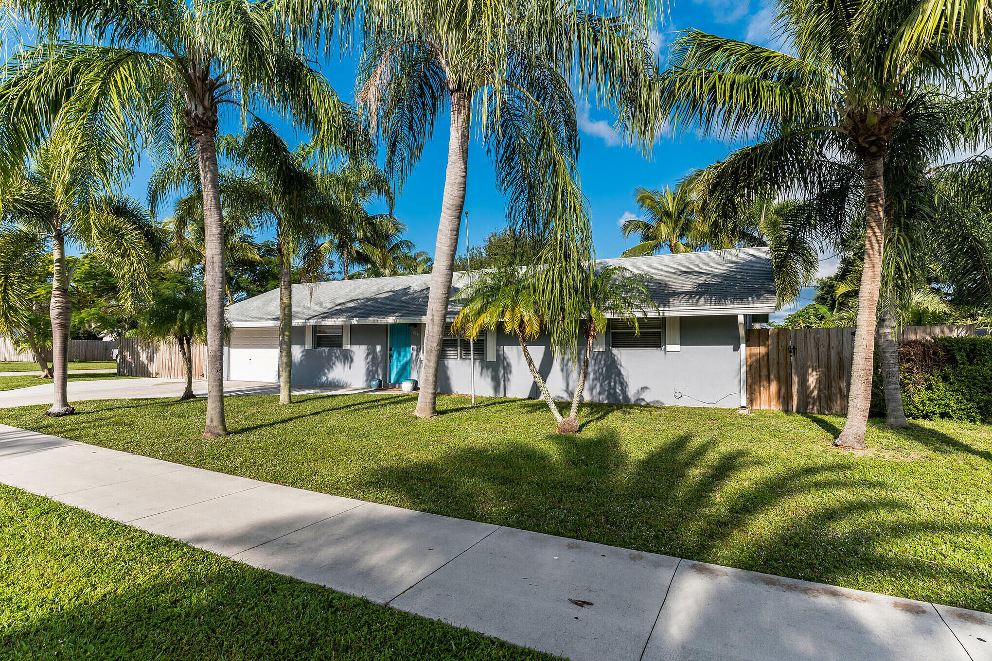 Single-story gray house with a turquoise door, palm trees, and a broad green lawn.
