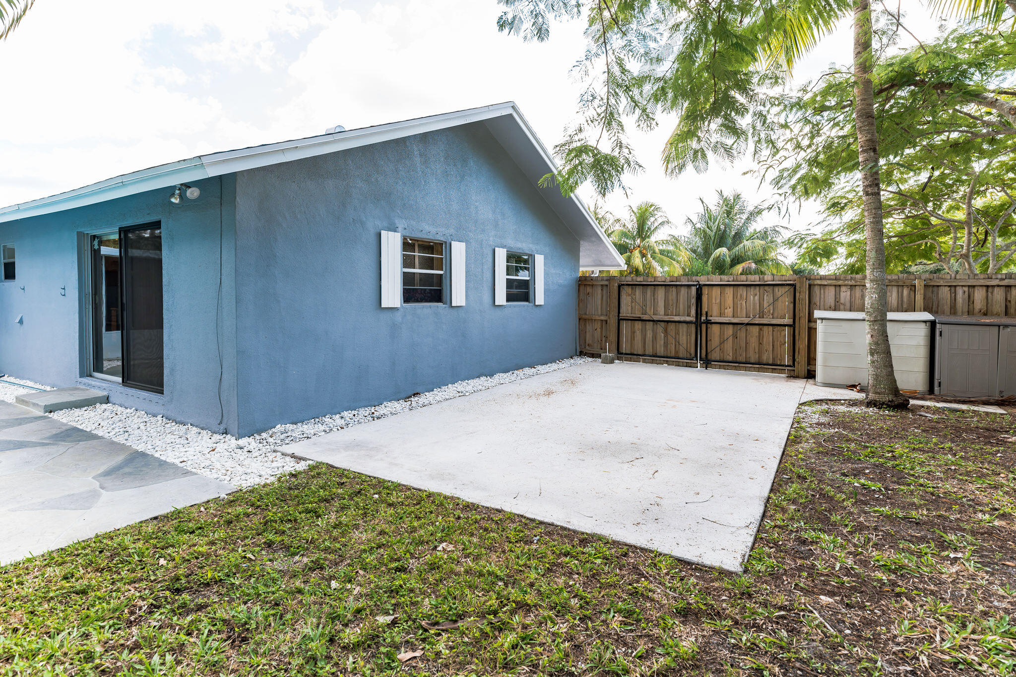 Blue home’s side patio with concrete paving, sliding door, wooden gate, and palm trees.