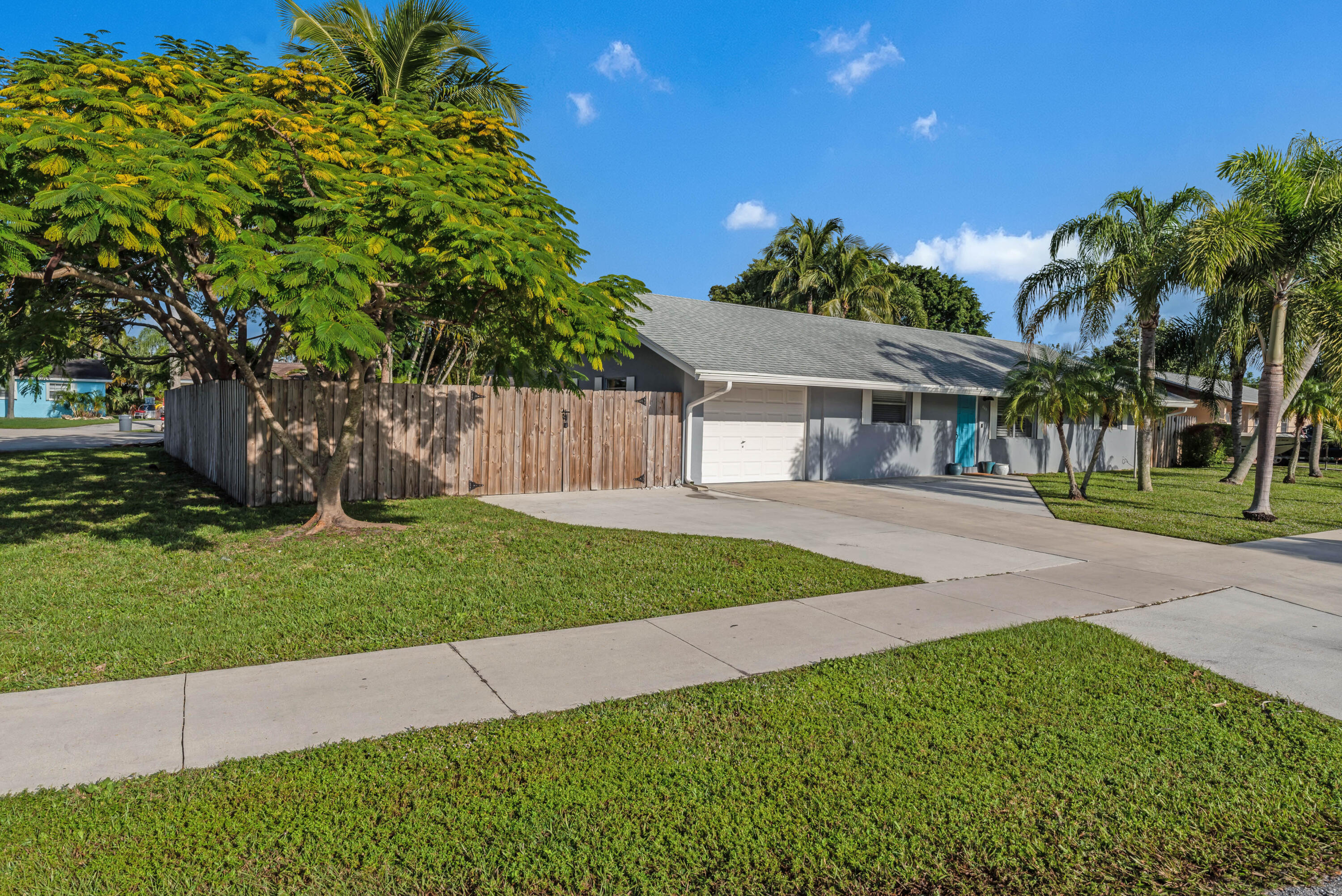 Single-story gray house with a white garage, turquoise front door, wooden fence, and palm-lined lawn.