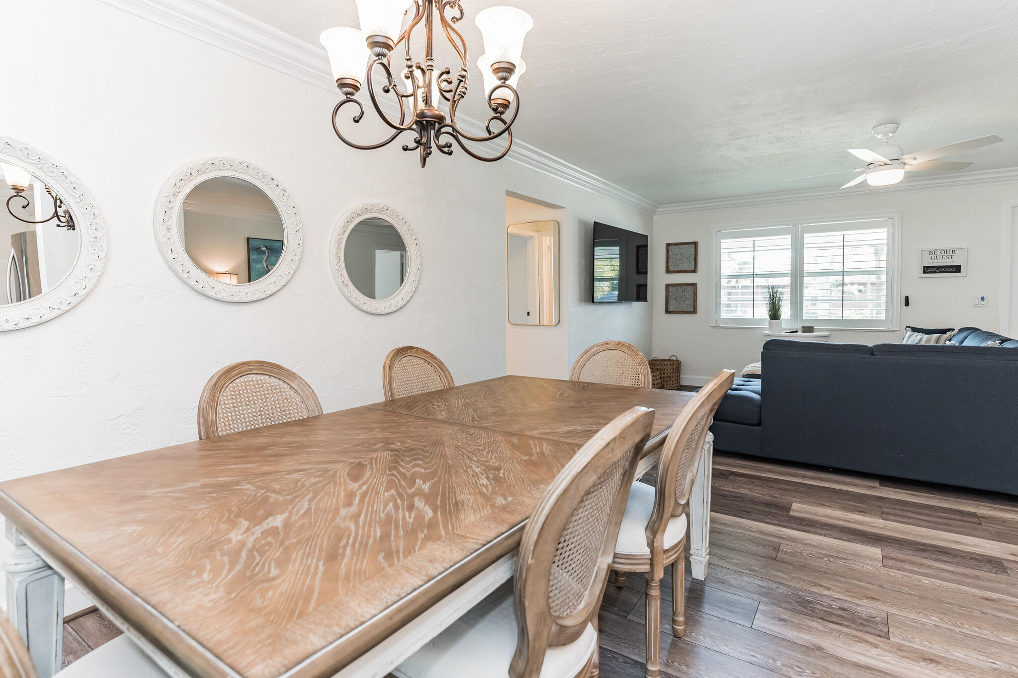 Dining room with a large wood table and woven chairs, opening onto a living room with a dark blue sofa.