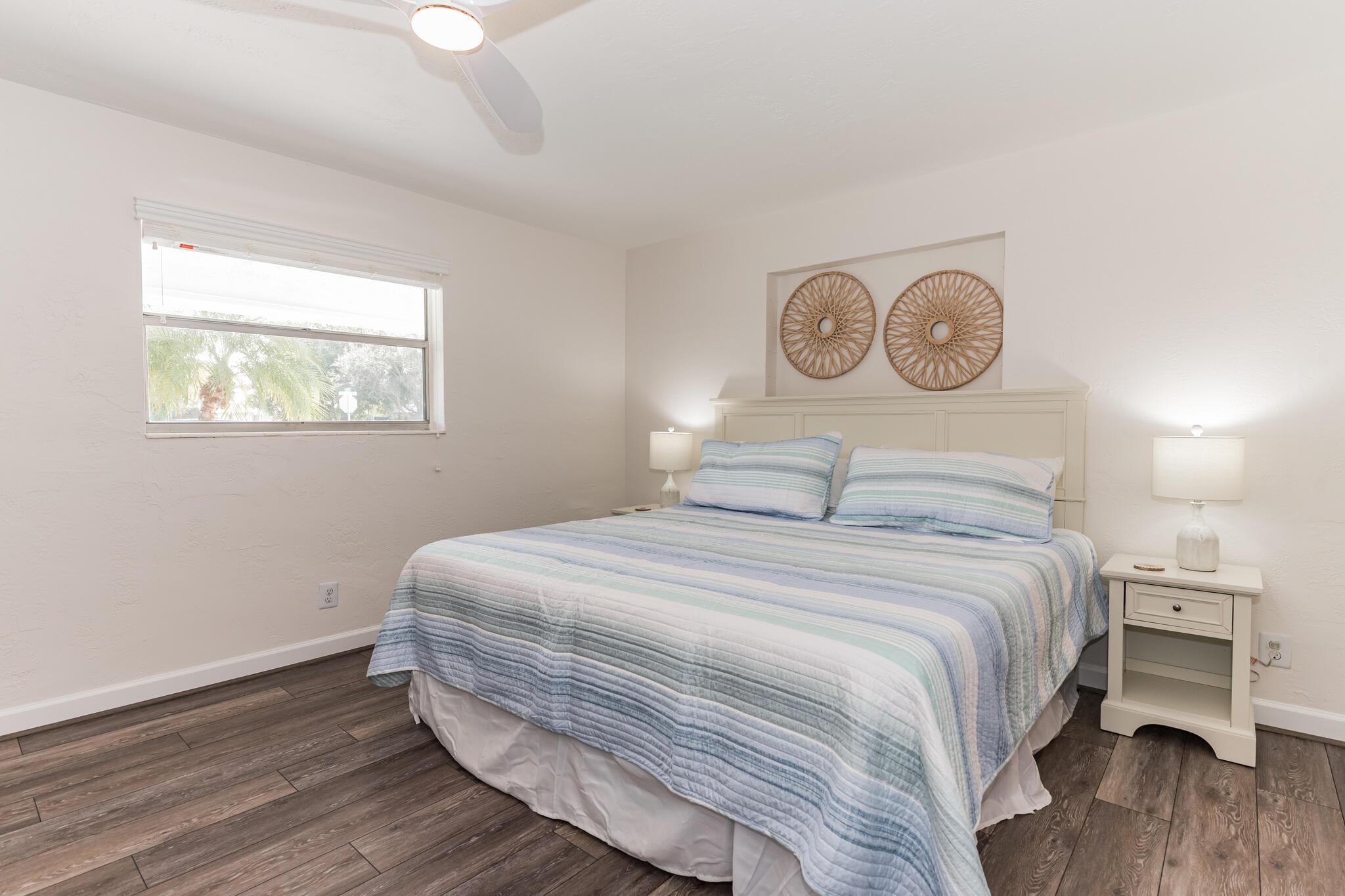 Bedroom with blue-and-white striped bedding, a white headboard, and a window overlooking palm trees.