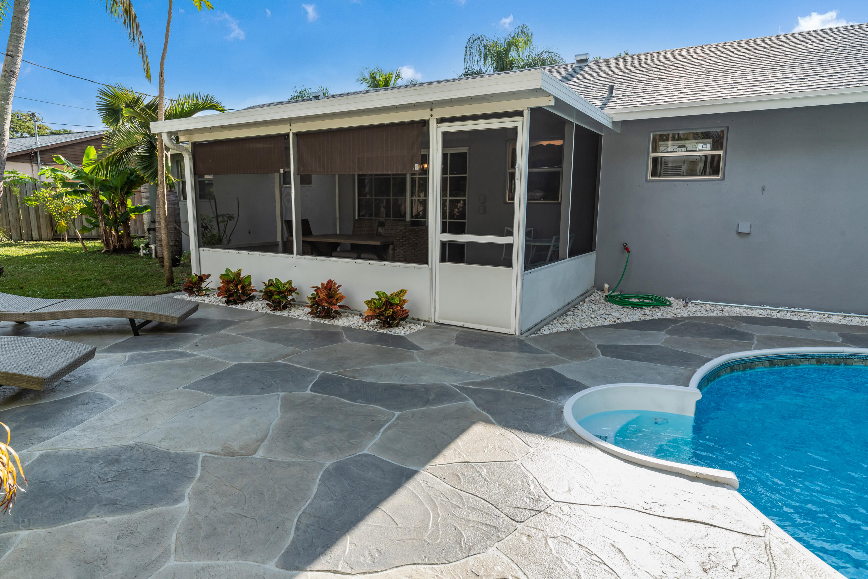 Backyard pool beside a stone patio and screened porch with tropical landscaping.