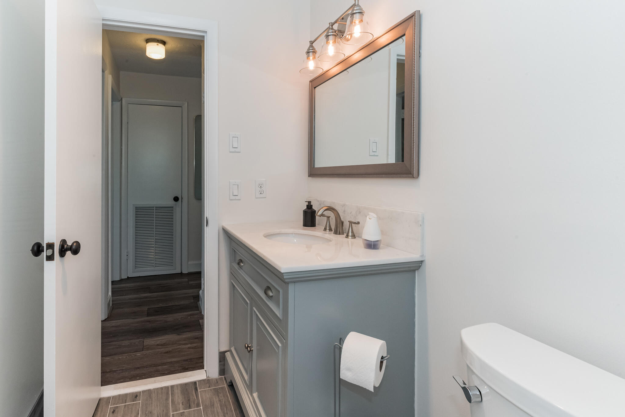 Bathroom with a gray vanity, framed mirror, and open doorway to a dark-floored adjoining space.