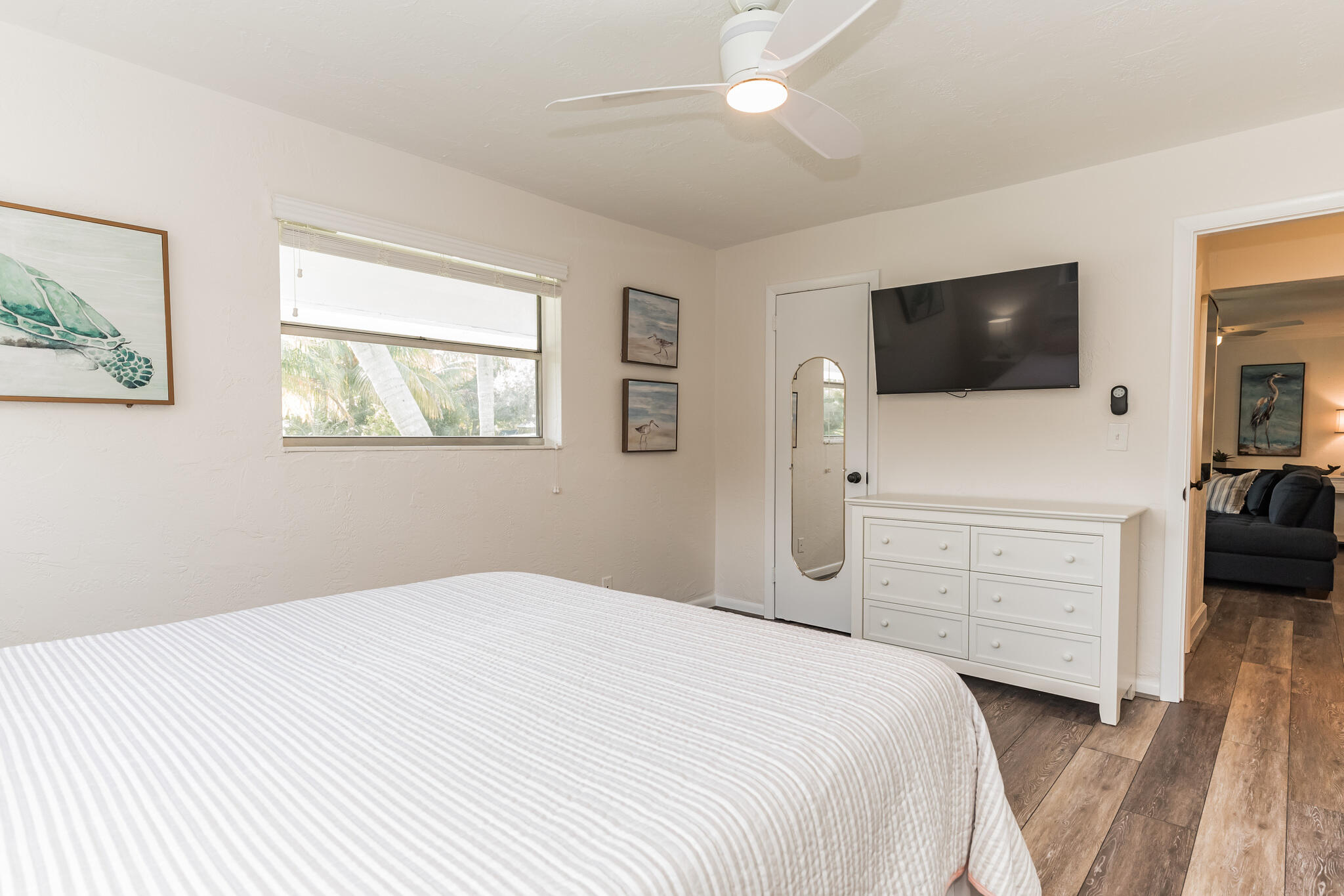 Bedroom with a striped white bed, dresser, wall-mounted TV, and a window overlooking palm trees.