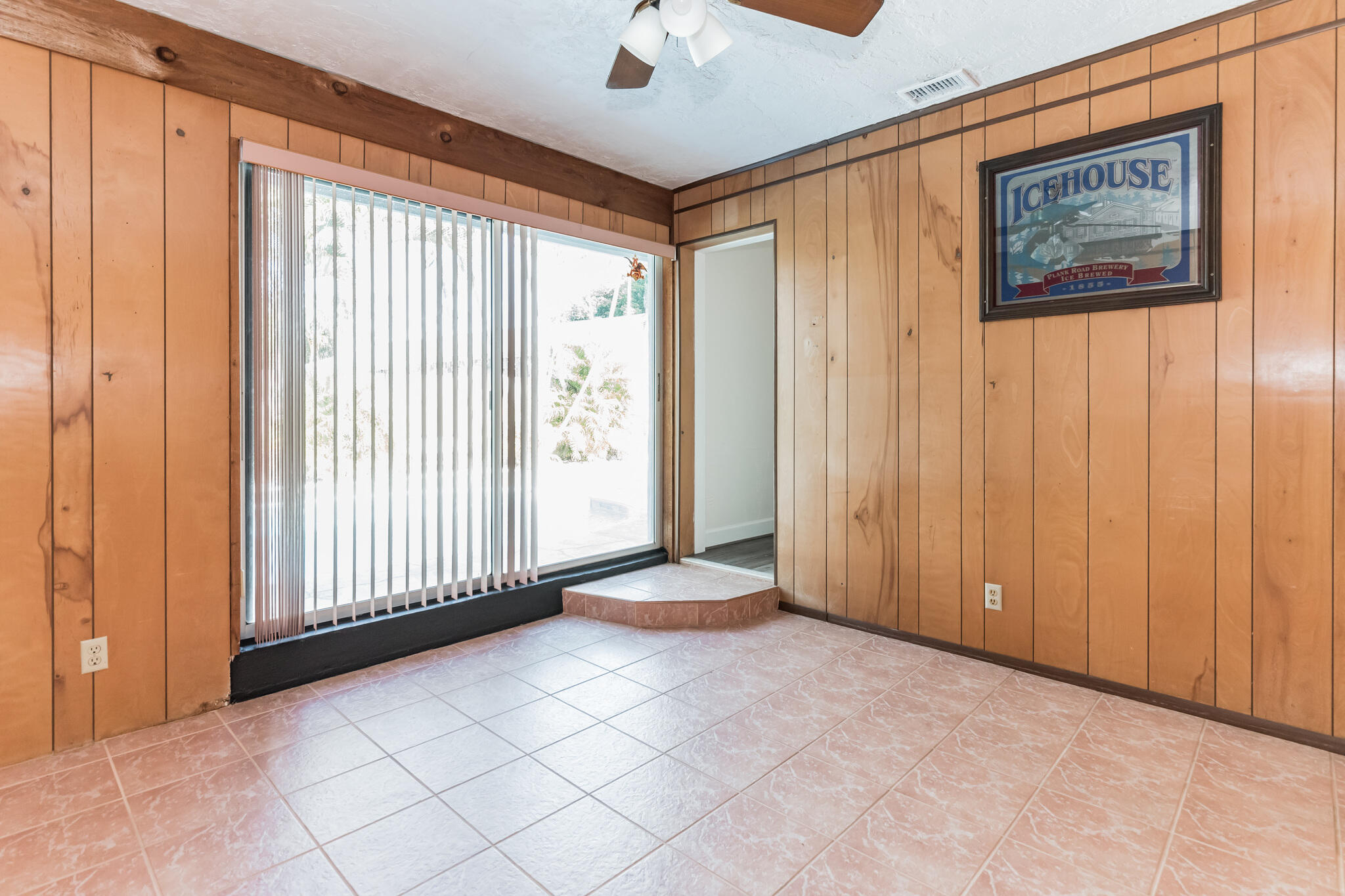 Empty wood-paneled room with pink tile flooring, vertical blinds, and a framed Icehouse poster.