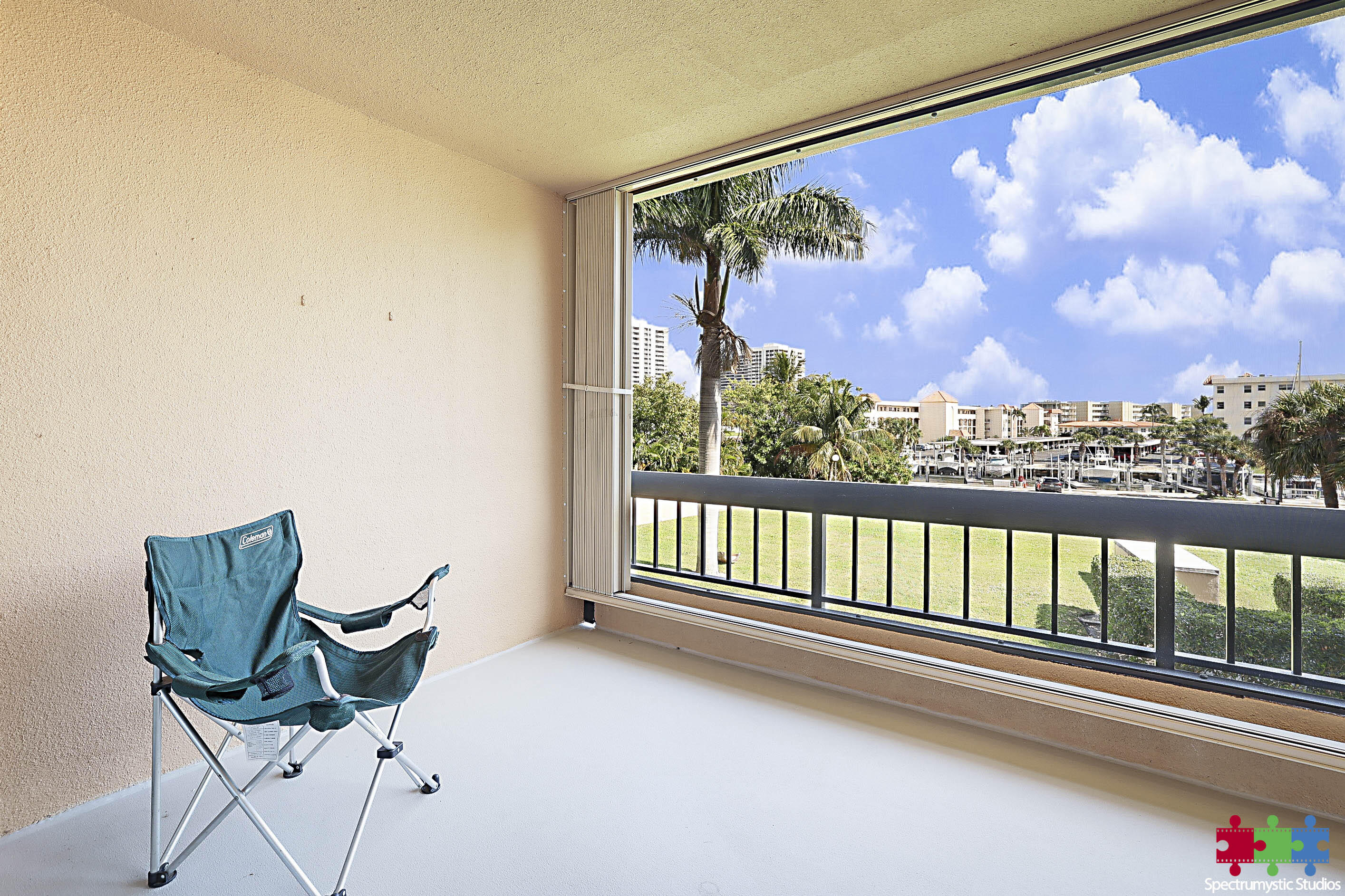 Covered condo balcony with a teal folding chair overlooking a marina and palm-lined waterfront.