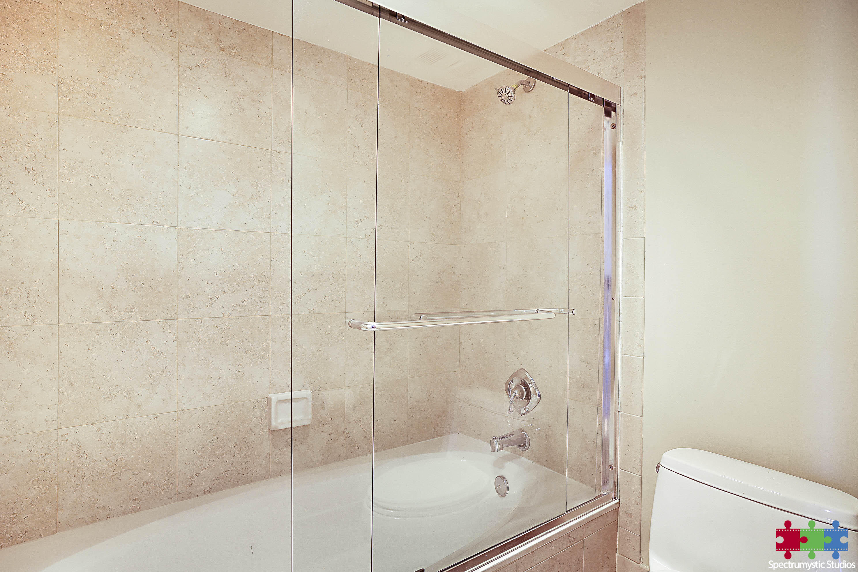 Beige stone-tiled bathroom with a glass-enclosed bathtub, chrome fixtures, grab bar, and adjacent toilet.