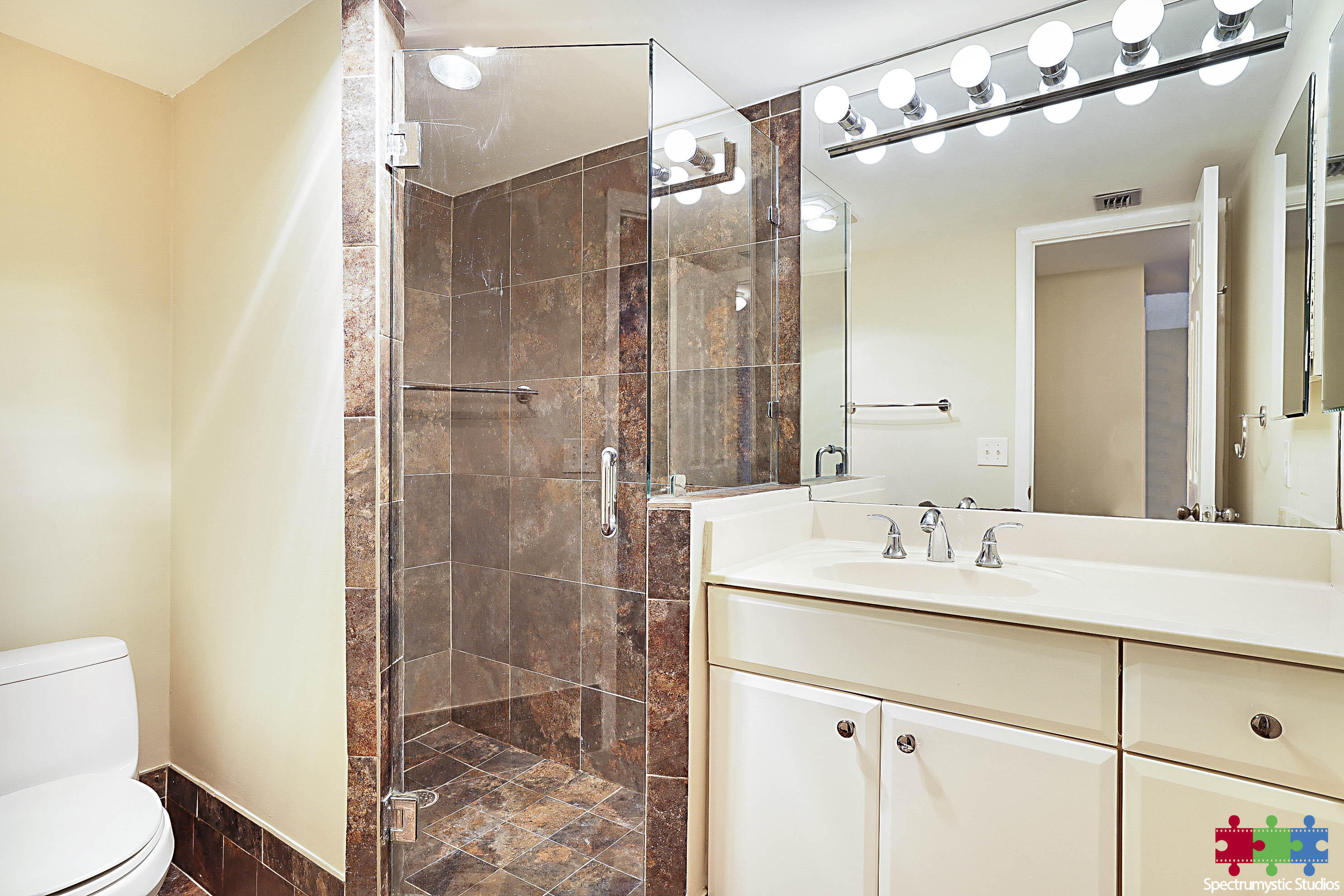 Bathroom with a glass-enclosed brown-tiled shower, white vanity, large mirror, and toilet.