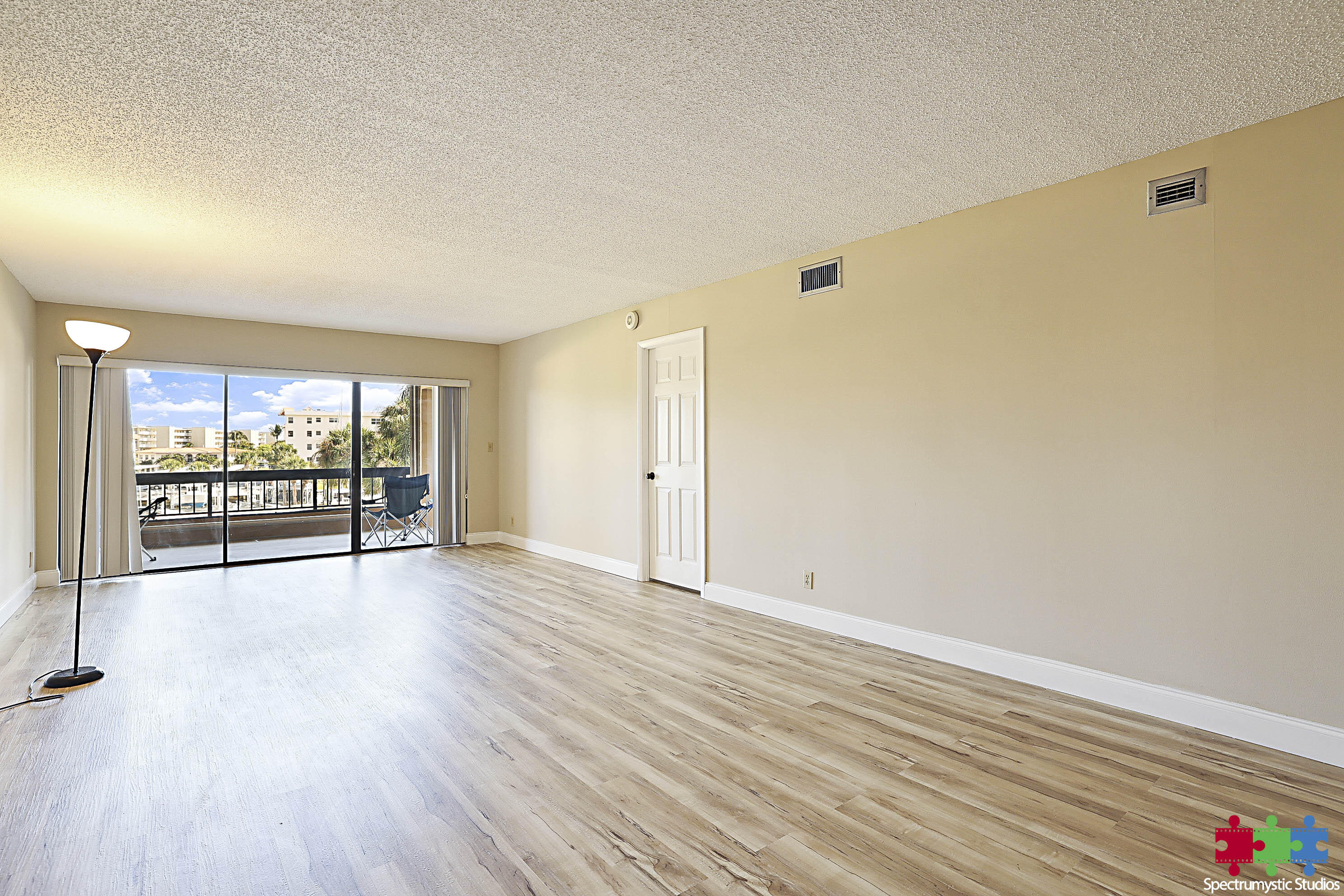 Empty condo living room with light wood-look floors and sliding doors to a balcony overlooking nearby buildings.