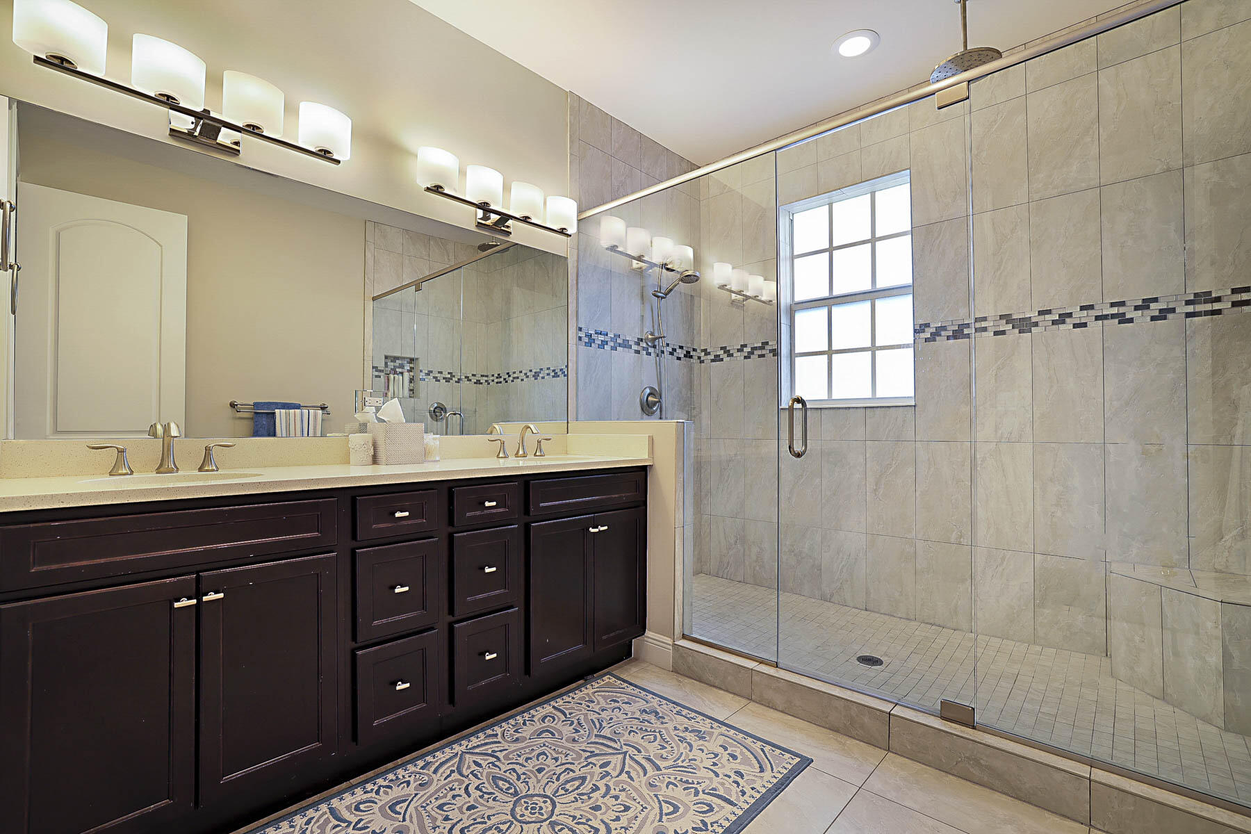 Bathroom with a dark double vanity and a glass-enclosed tiled shower beside a window.