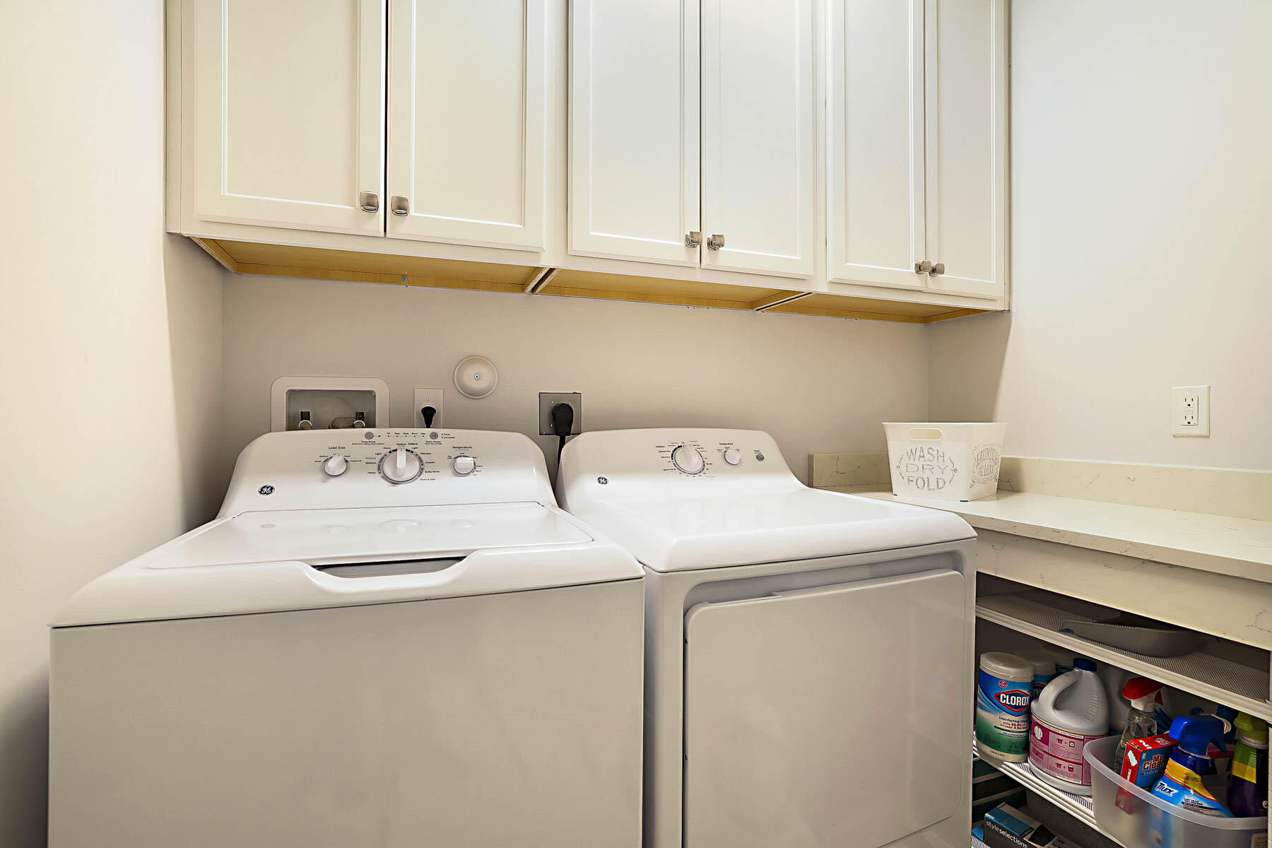 Laundry room with white washer and dryer, upper cabinets, and cleaning supplies on open shelves.