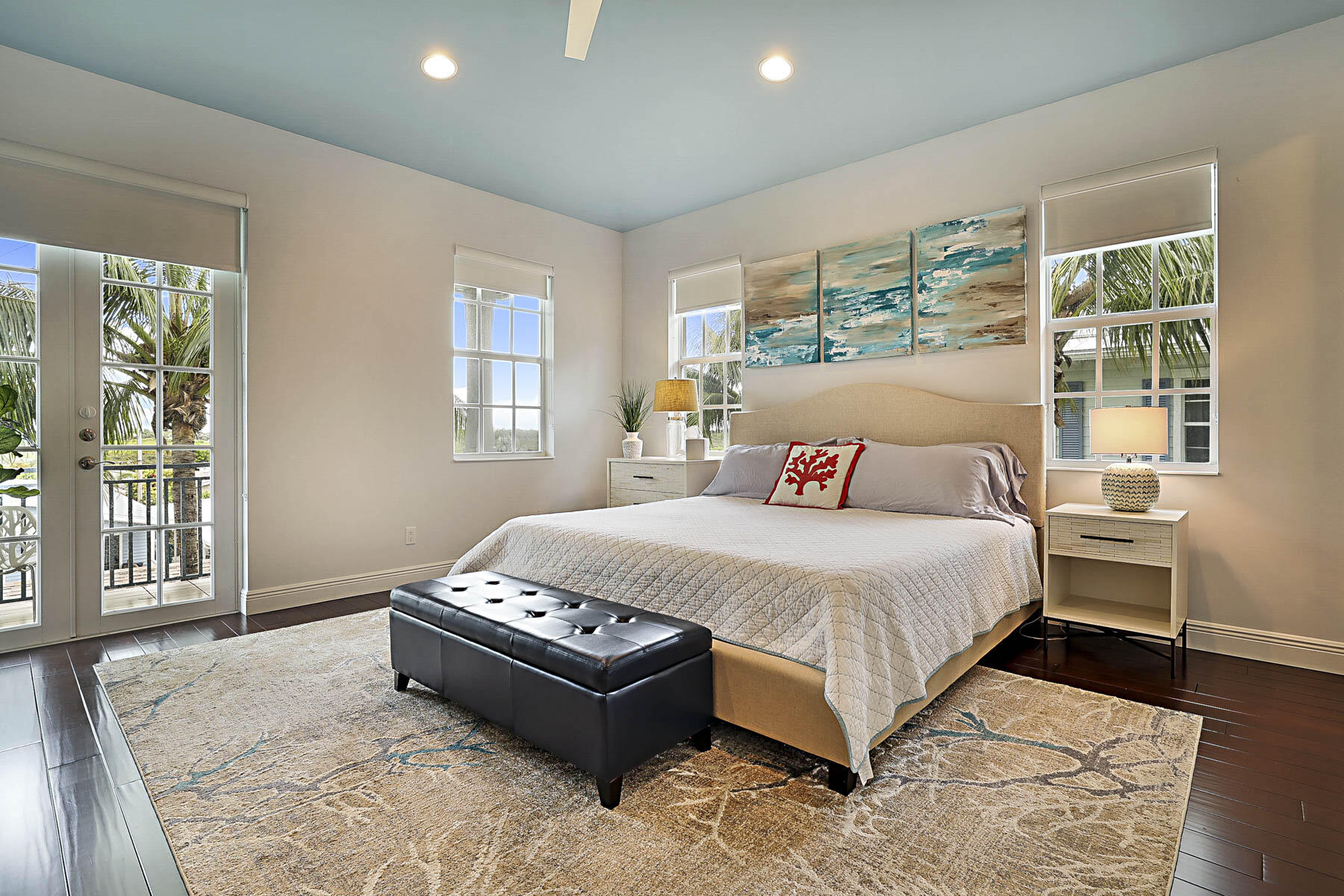 Furnished bedroom with a large bed, black bench, coastal artwork, and windows overlooking palm trees.