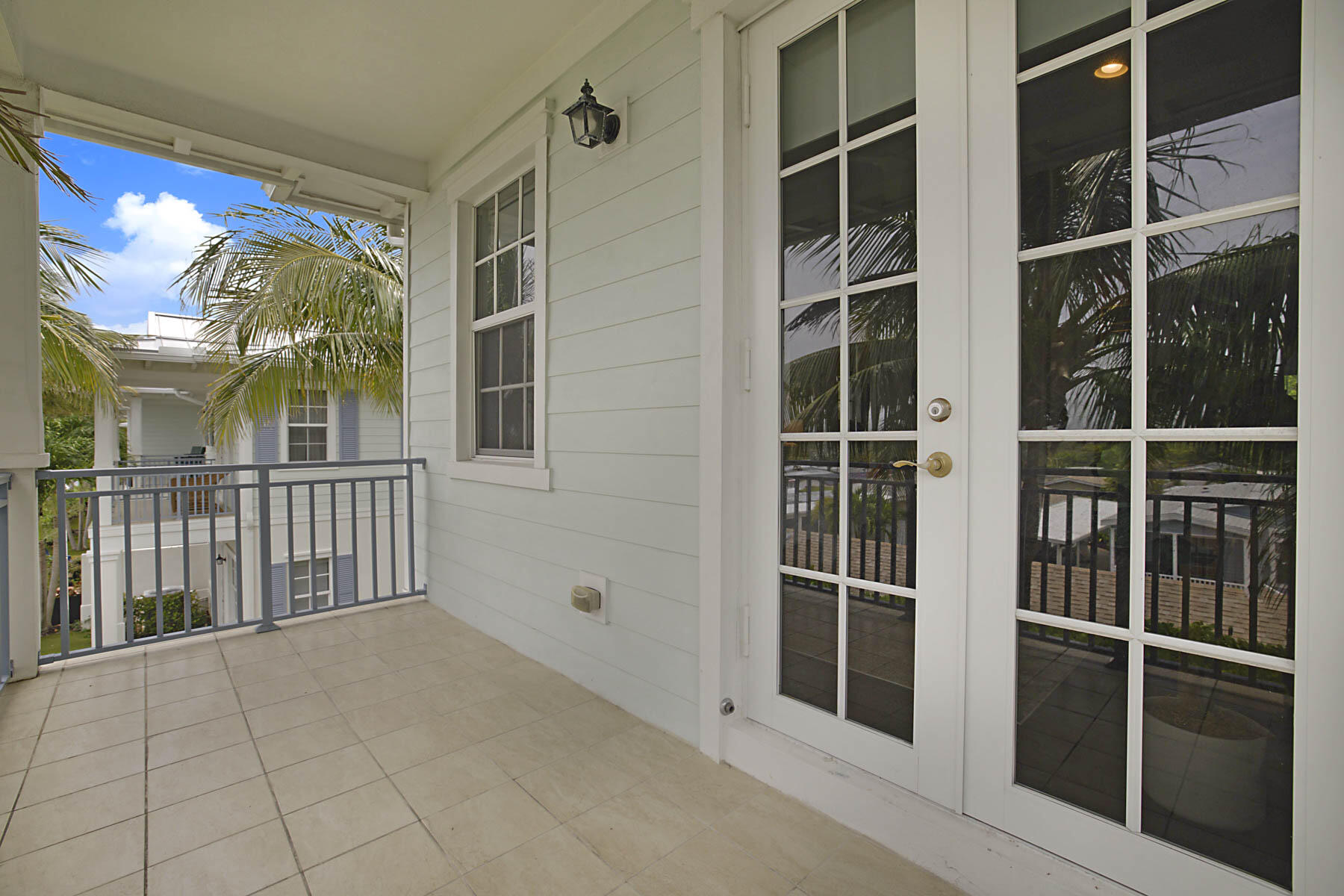 Covered tiled balcony with white French doors, gray railing, and palm trees.