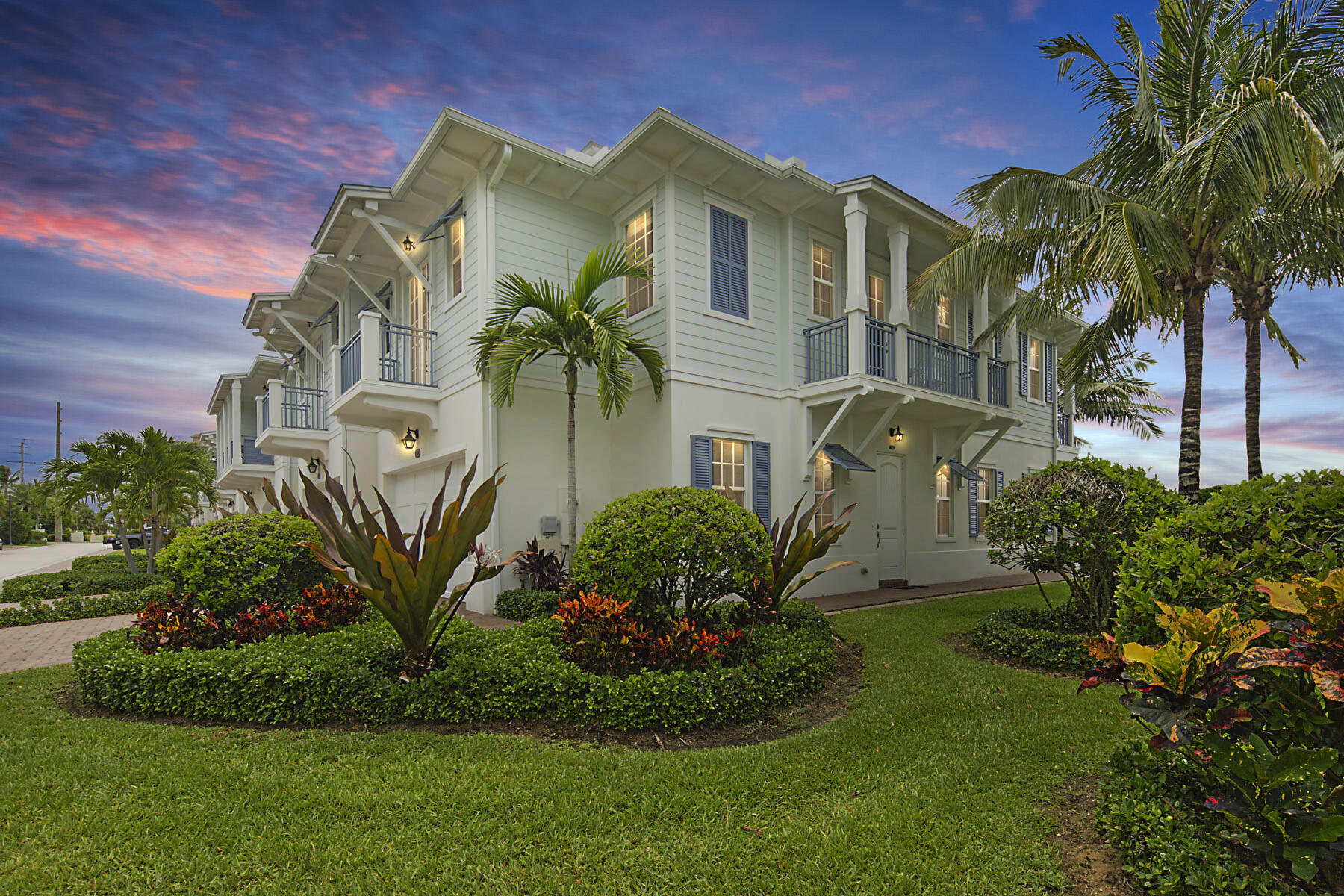 White coastal-style residence with blue-shuttered windows, balconies, and tropical landscaping at dusk.