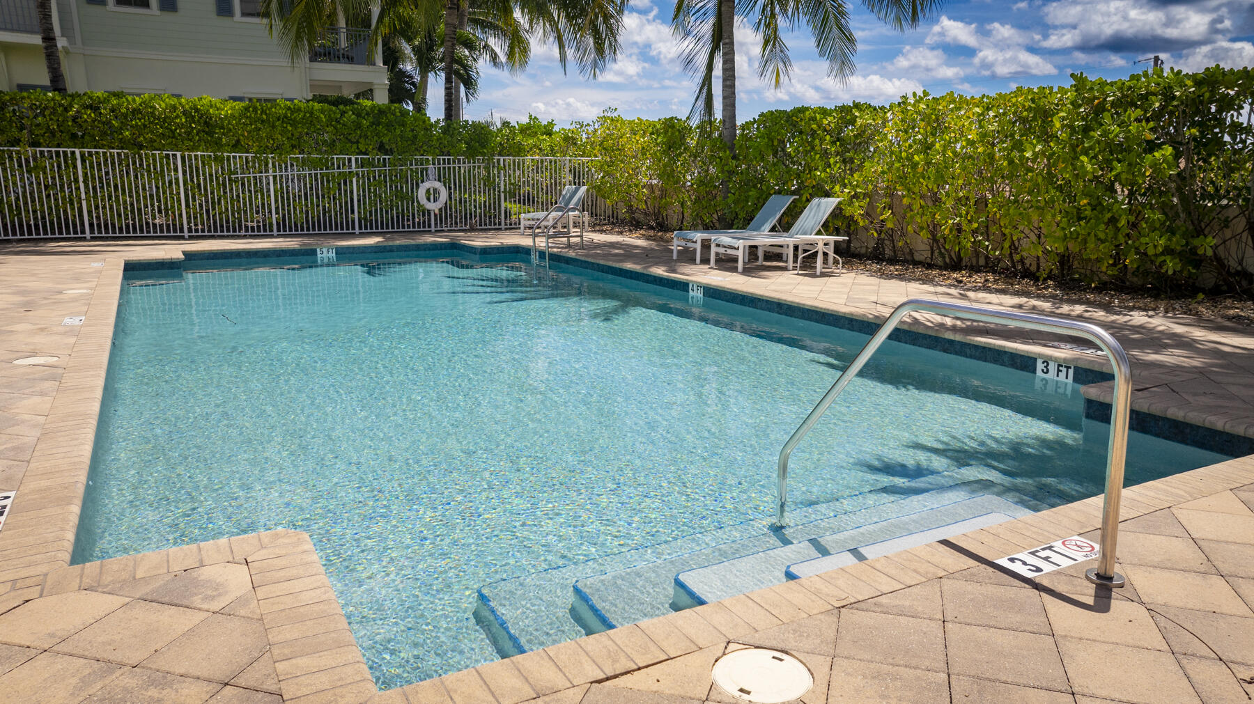 Outdoor swimming pool with shallow entry steps, metal handrails, and two lounge chairs beside a hedge.