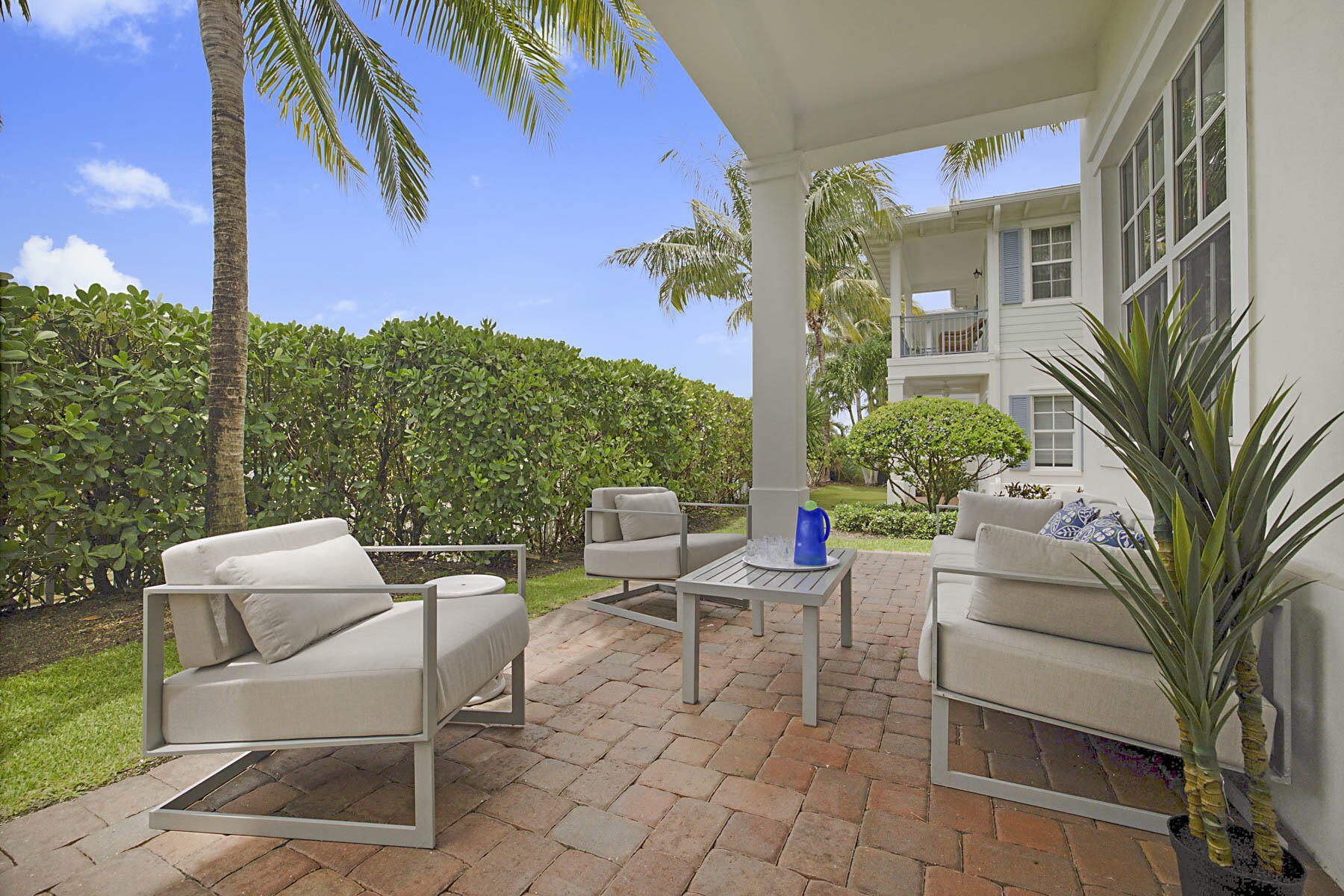 Covered brick patio with lounge seating, palm trees, and a hedge beside a white house.