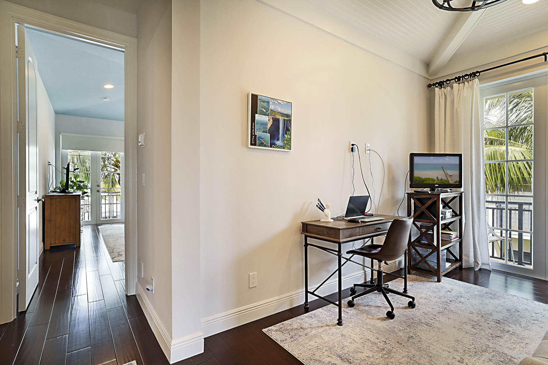 Furnished home office with a wooden desk and chair beside palm-framed balcony doors.