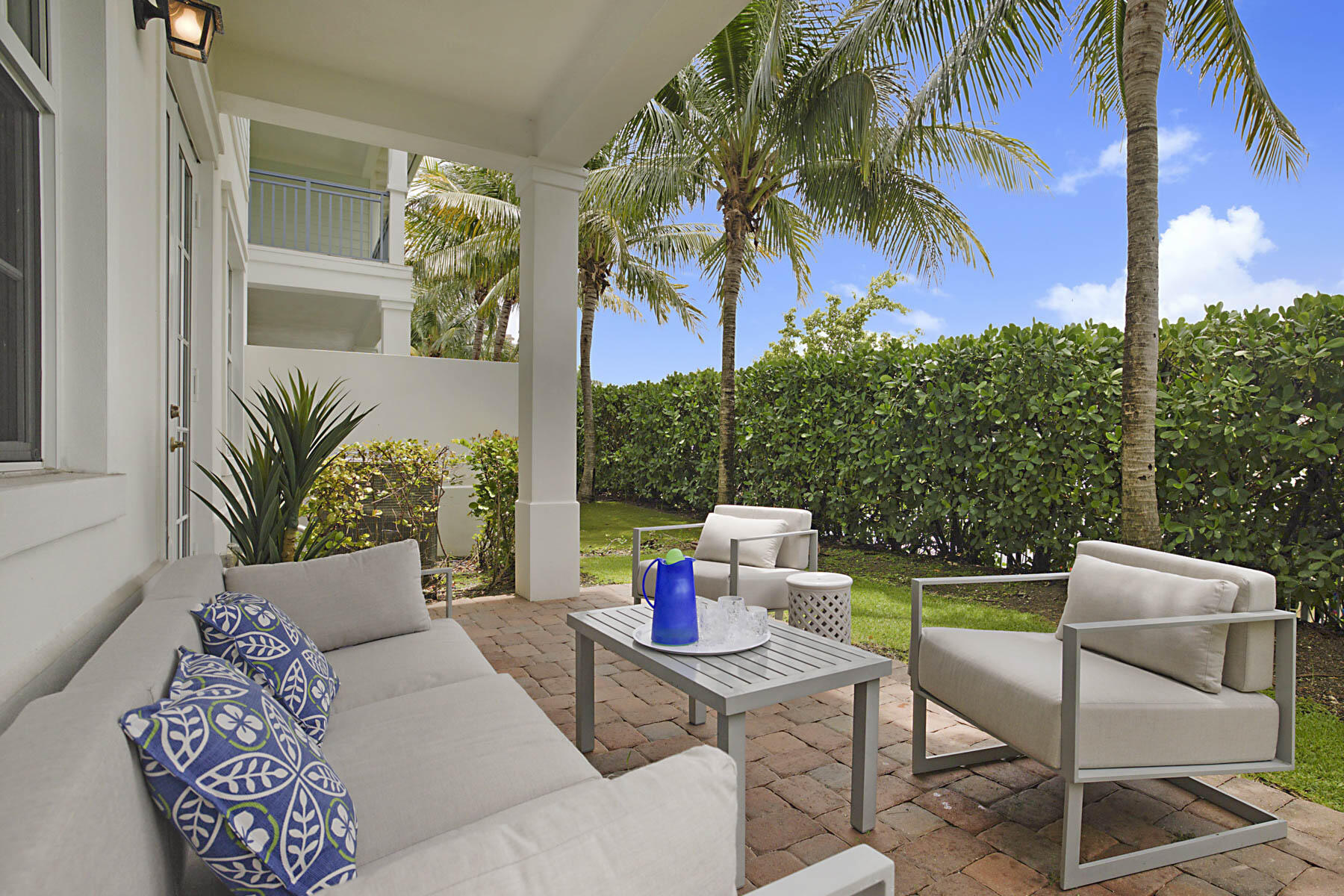 Covered patio with cushioned seating, a coffee table, and palm trees beside a tall hedge.