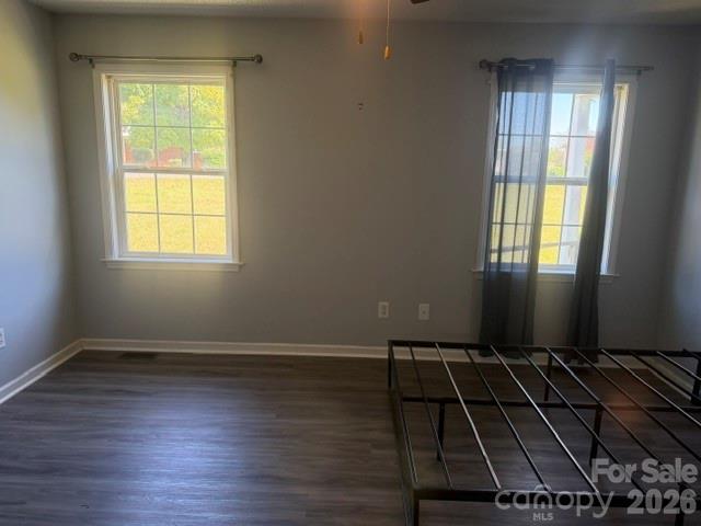 Empty bedroom with two curtained windows, dark flooring, and part of a metal bed frame.