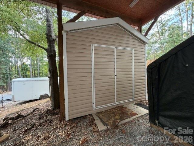Beige storage shed with sliding doors beneath a covered structure, with woods and a white trailer nearby.