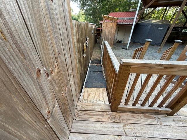 Wooden deck walkway beside a tall privacy fence, overlooking a gravel yard and covered structures.