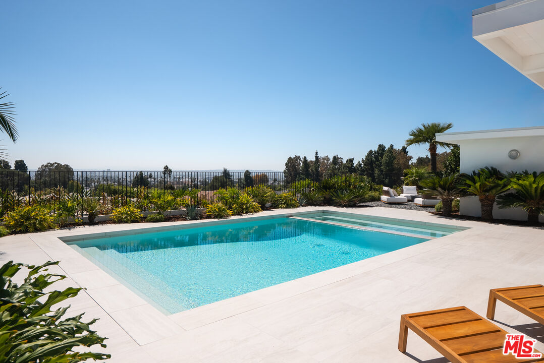 Swimming pool on a pale stone terrace with tropical landscaping and a distant city view.