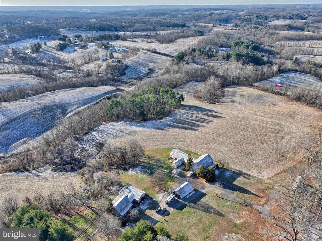 Aerial view of rural buildings and expansive fields with patches of snow and wooded edges.