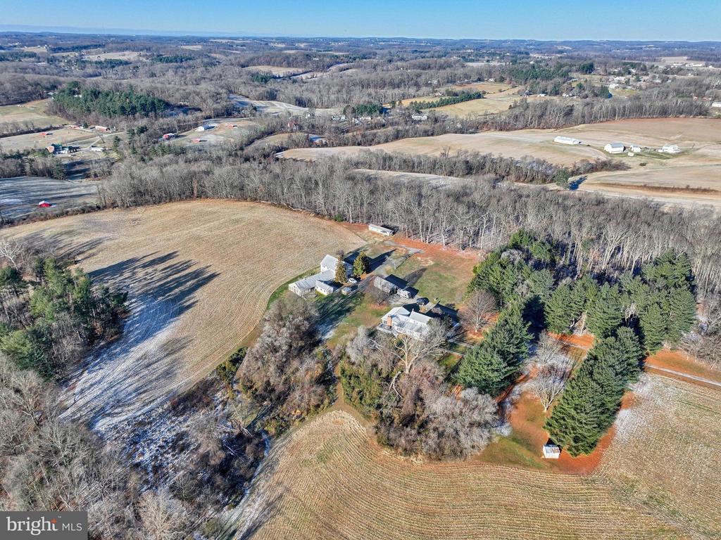 Aerial view of a rural home, outbuildings, open fields, and surrounding woods.