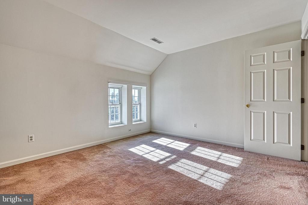 Vacant bedroom with beige carpet, two windows, and sunlight casting shadows across the floor.