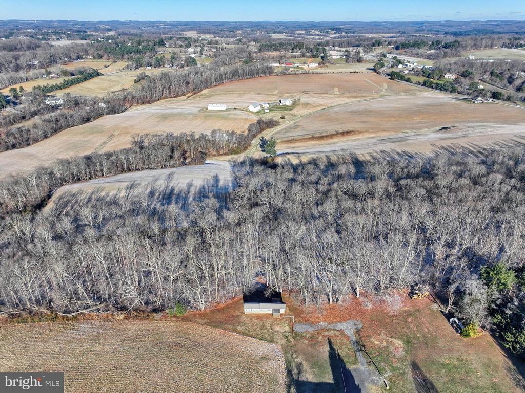Aerial view of a rural home beside leafless woods, with open fields and rolling countryside beyond.