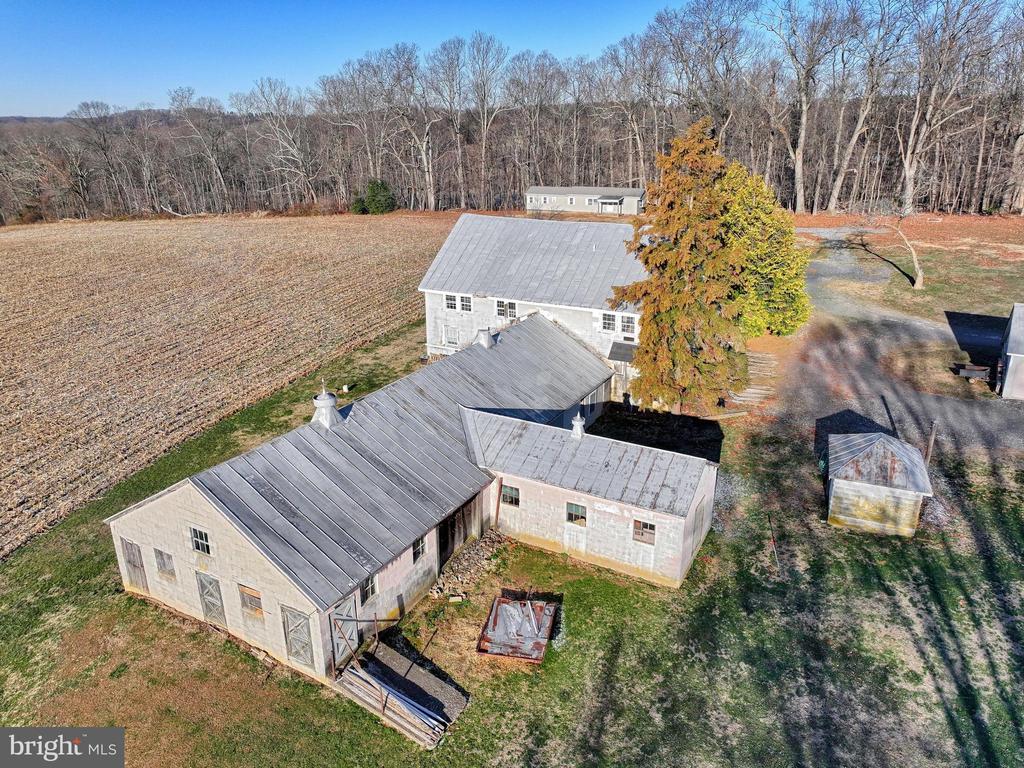 Aerial view of rural outbuildings beside a field, with a small house and wooded tree line behind them.