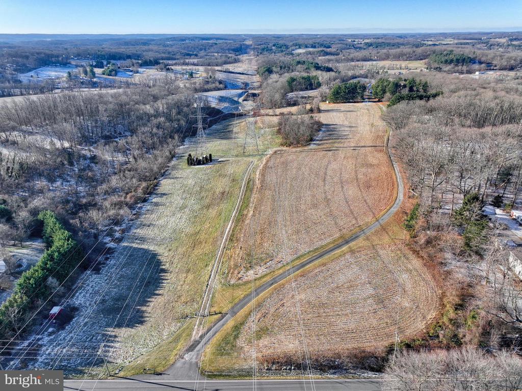 Aerial view of rural fields and wooded land with a curving access road and utility lines.