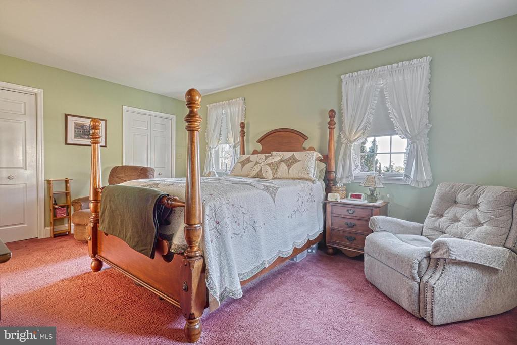 Bedroom with a wooden four-poster bed, white bedding, and an upholstered armchair beside curtained windows.
