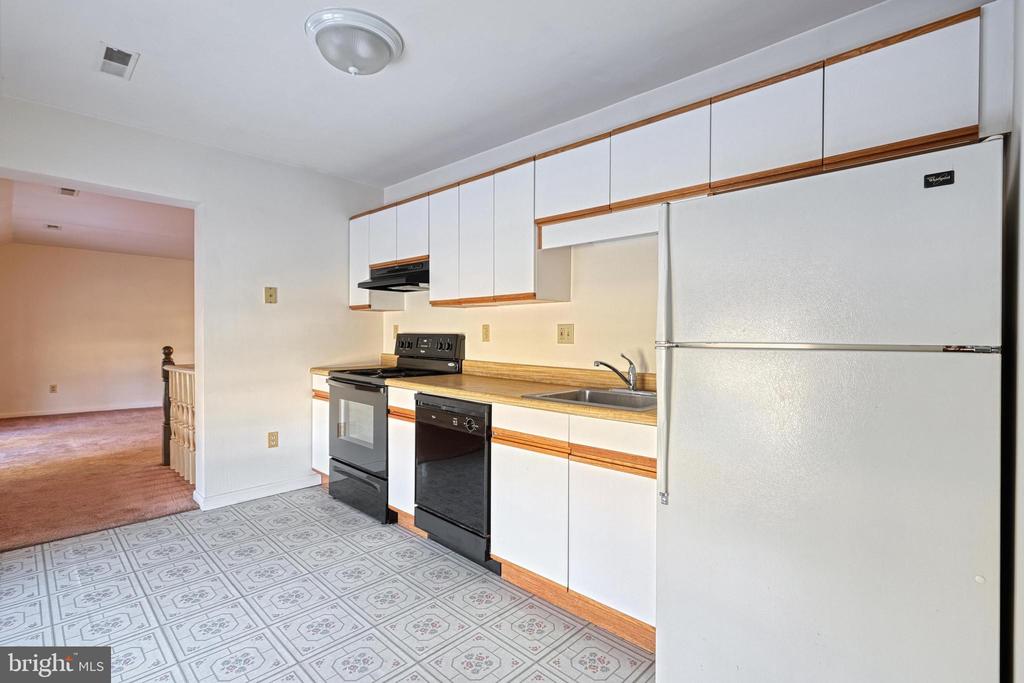 Kitchen with white, wood-trimmed cabinets, black appliances, patterned vinyl floor, and doorway to a carpeted room.