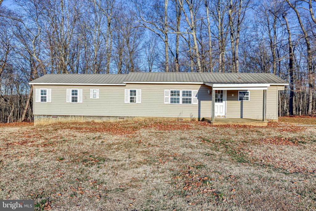 Single-story beige home with a metal roof and covered entrance, bordered by leafless woods.