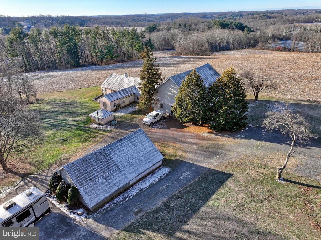 Aerial view of a rural home and outbuildings beside open fields and wooded hills.