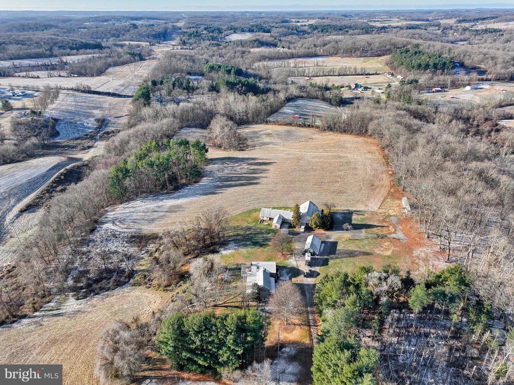 Aerial view of a broad rural field bordered by woods, with several outbuildings near the driveway.
