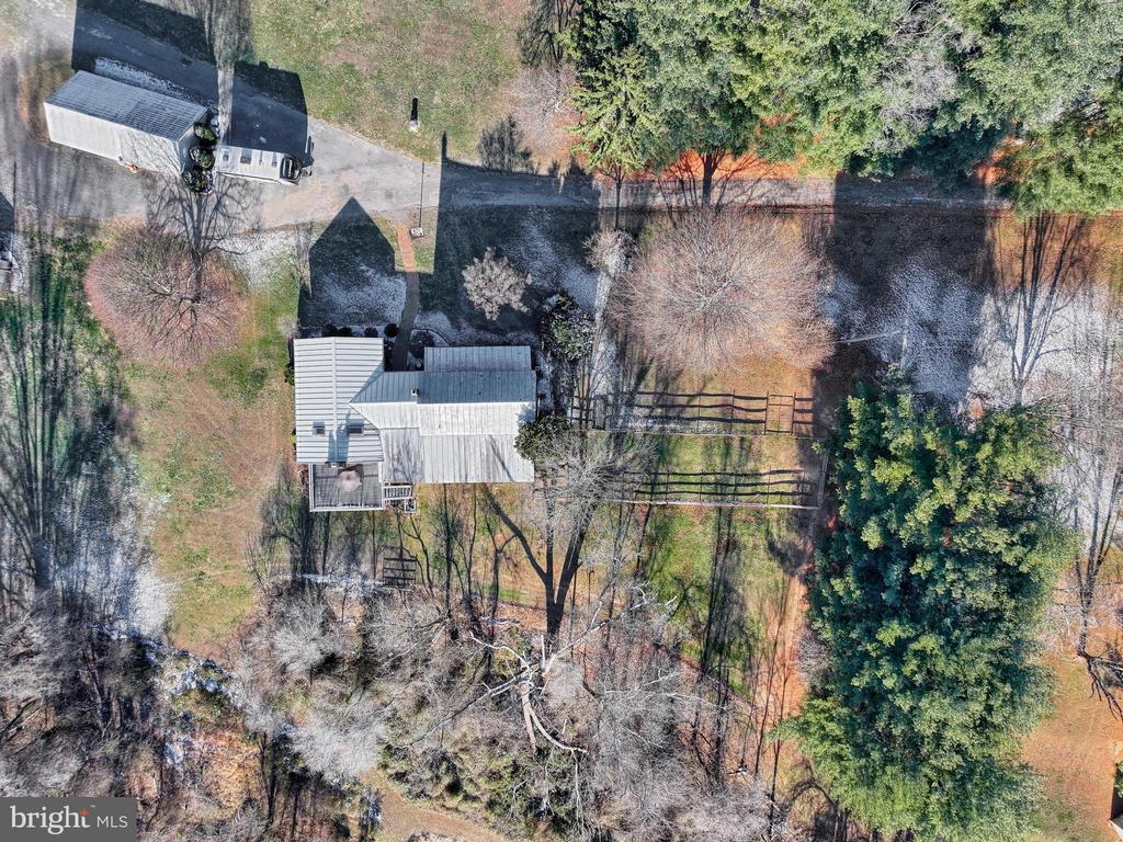 Aerial view of a house with a detached outbuilding, driveway, fenced yard, and surrounding trees.