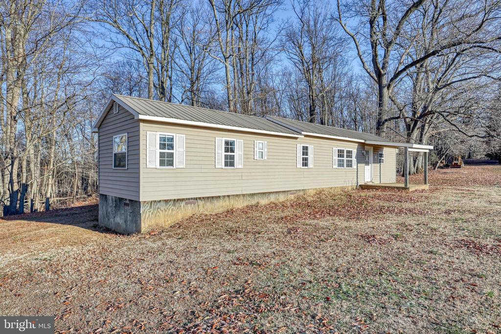 Single-story beige home with a gray metal roof, covered entrance, and wooded surroundings.